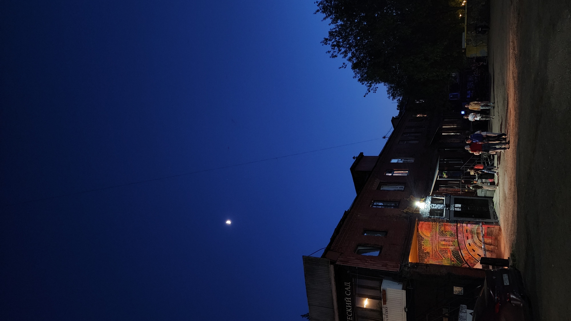 At night, the sky is a deep blue with a thin crescent moon visible. A brick building stands alongside a road where a group of people congregate near an entrance featuring street art. The building's windows are dark except for one illuminated on the lower level. The setting is Moscow, Russia.