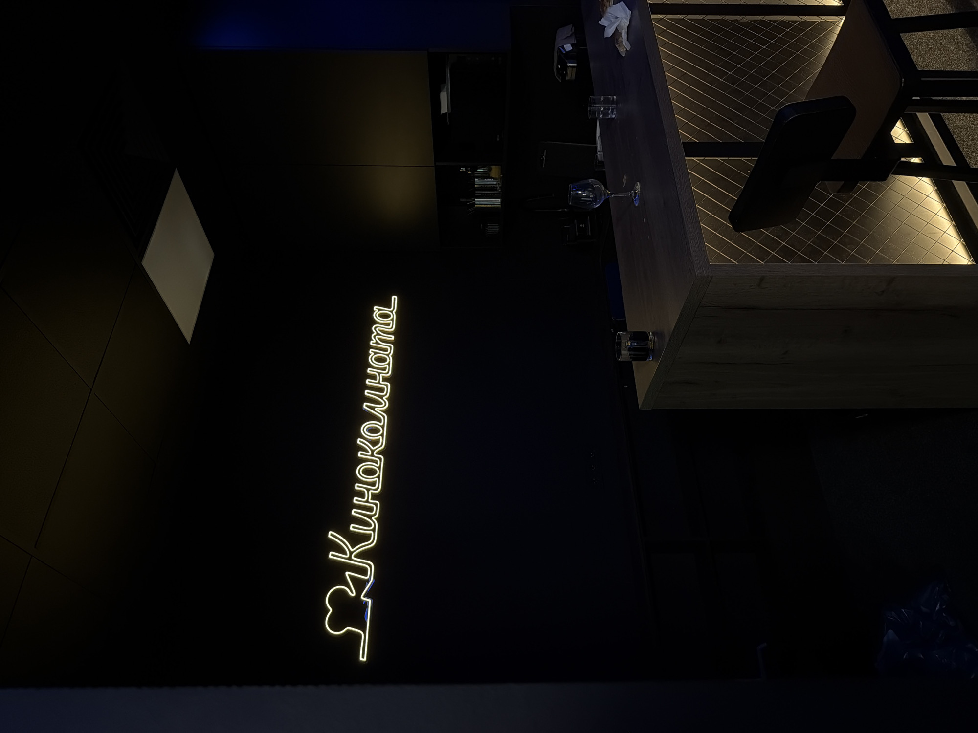 The image depicts a dimly lit bar area with a neon sign on the wall that reads "Киноkomnata" in yellow lettering, accompanied by a heart-shaped film reel icon. A wooden bar counter is visible in the foreground, with a few glasses and a crumpled napkin on its surface. A single bar stool with a dark wooden seat and back is positioned in front of the counter. The bar itself features a decorative front with a diamond pattern and integrated lighting. In the background, there are shelves with bottles and other bar items, as well as a mirror. The overall ambiance is dark and moody, suggesting a late-night or evening setting. The location is identified as Unknown, Unknown.