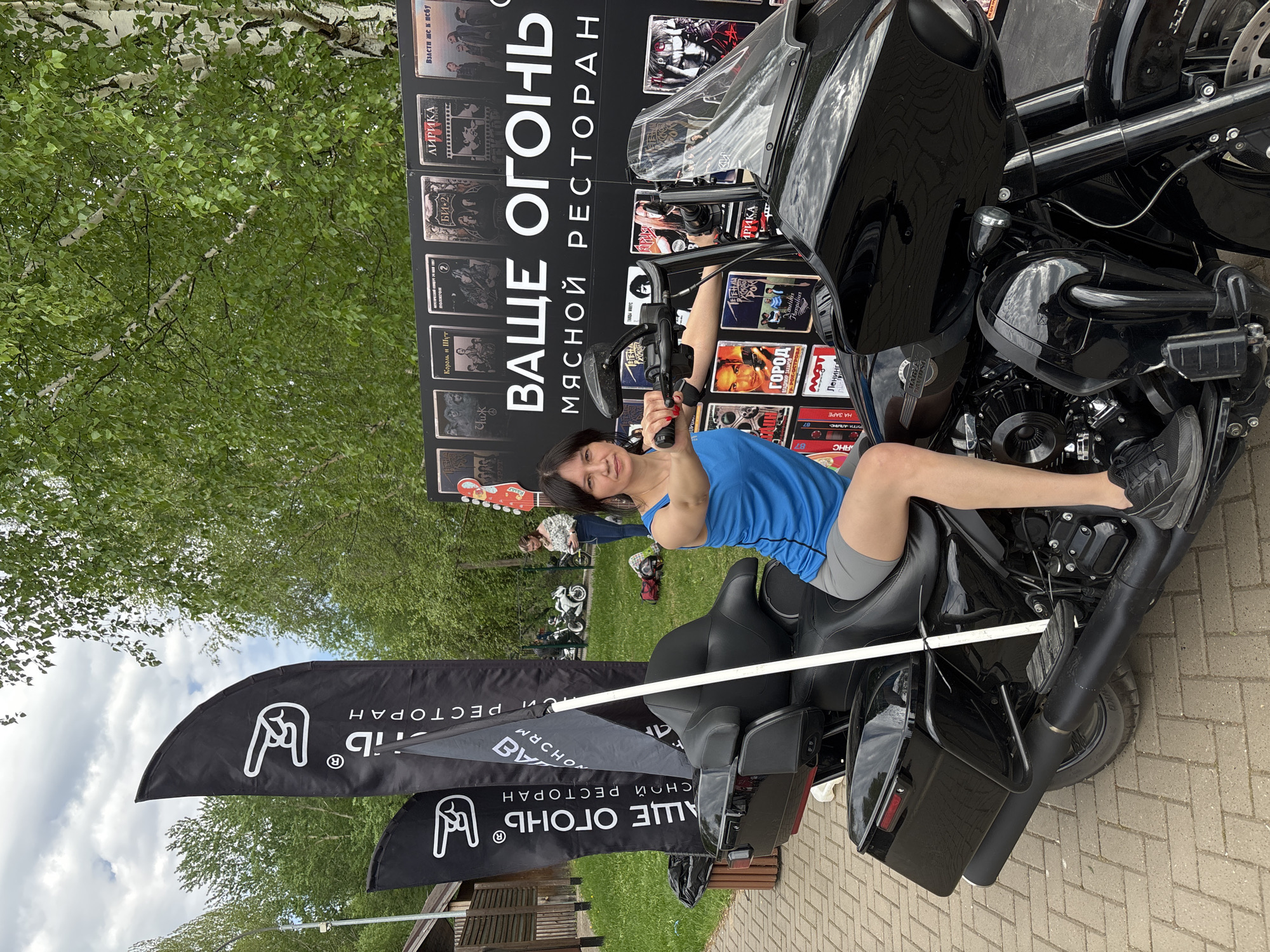 Here is a detailed description of the image:
**Scene:** The image appears to be taken outdoors, possibly in a parking area or event space, given the paved surface and the presence of grassy areas and trees in the background.
**Subjects:** The main subject is a woman posing on a large, black motorcycle. She is wearing a blue tank top and grey shorts. Behind her is a large sign with Cyrillic text and images that appear to be album covers, advertising a restaurant called "ВАЩЕ ОГОНЬ" (YOUR FIRE) with the tagline "МЯСНОЙ РЕСТОРАН" (MEAT RESTAURANT). There are black flags with a white hand gesture logo and the same Cyrillic text as the sign. Other motorcycles and people can be seen blurred in the background.
**Objects:** The primary objects are the motorcycle, the restaurant advertising sign, and the flags. Additional objects include the paved ground, trees, and other distant motorcycles and people.
**Actions/Interactions:** The woman is interacting with the motorcycle by sitting on it and posing for the camera. The motorcycle seems to be stationary. The environment suggests a gathering or event, possibly related to motorcycles and/or the advertised restaurant.