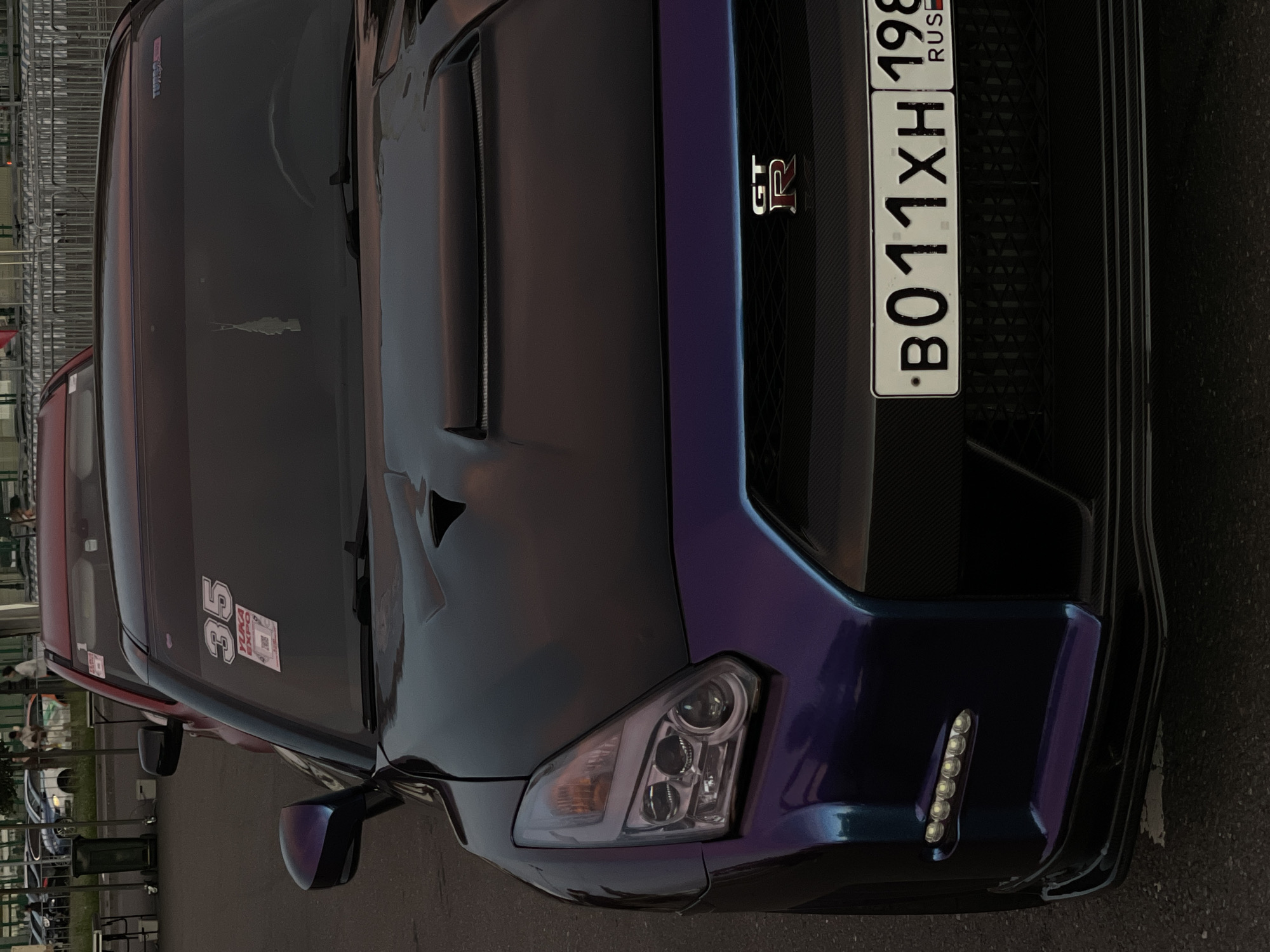 An exterior, eye-level, medium shot captures the front of a dark-colored Nissan GT-R. The car's hood displays a subtle purple and blue iridescent sheen, particularly on its fenders and hood scoop. The vehicle's headlights are complex, with multiple circular beams and a distinct amber turn signal. A red sticker with the number "35" and "YUKA EXPO" is affixed to the windshield on the driver's side. A small, white air freshener hangs from the rearview mirror. The front grille of the car is black and textured, featuring the "GT-R" emblem prominently. A Russian license plate, "B011XH 198," is mounted below the grille. The car is parked on an asphalt surface, likely a street or parking lot, with a hint of a metallic barrier in the background. Another vehicle, a red car, is partially visible behind the GT-R. The lighting suggests it is either early morning or late afternoon, casting soft shadows.