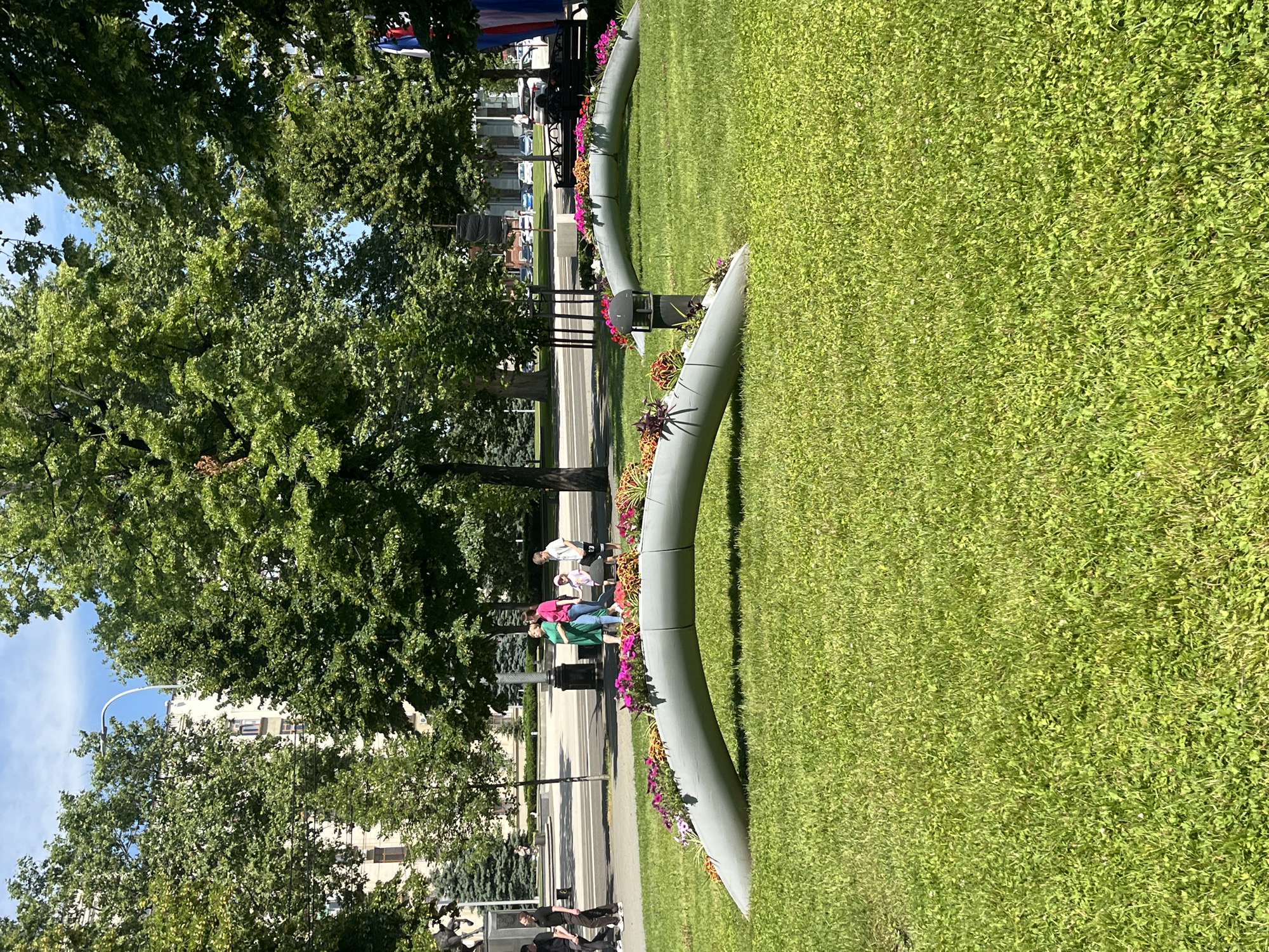 In Krasnodar, Russia, on a sunny day, two curved, gray planters filled with colorful flowers are situated on a vibrant green lawn. In the background, a paved walkway runs parallel to the lawn, with several people walking along it. Behind the walkway, trees line the path, and a building with a light-colored facade is visible. The sky above is a clear blue.