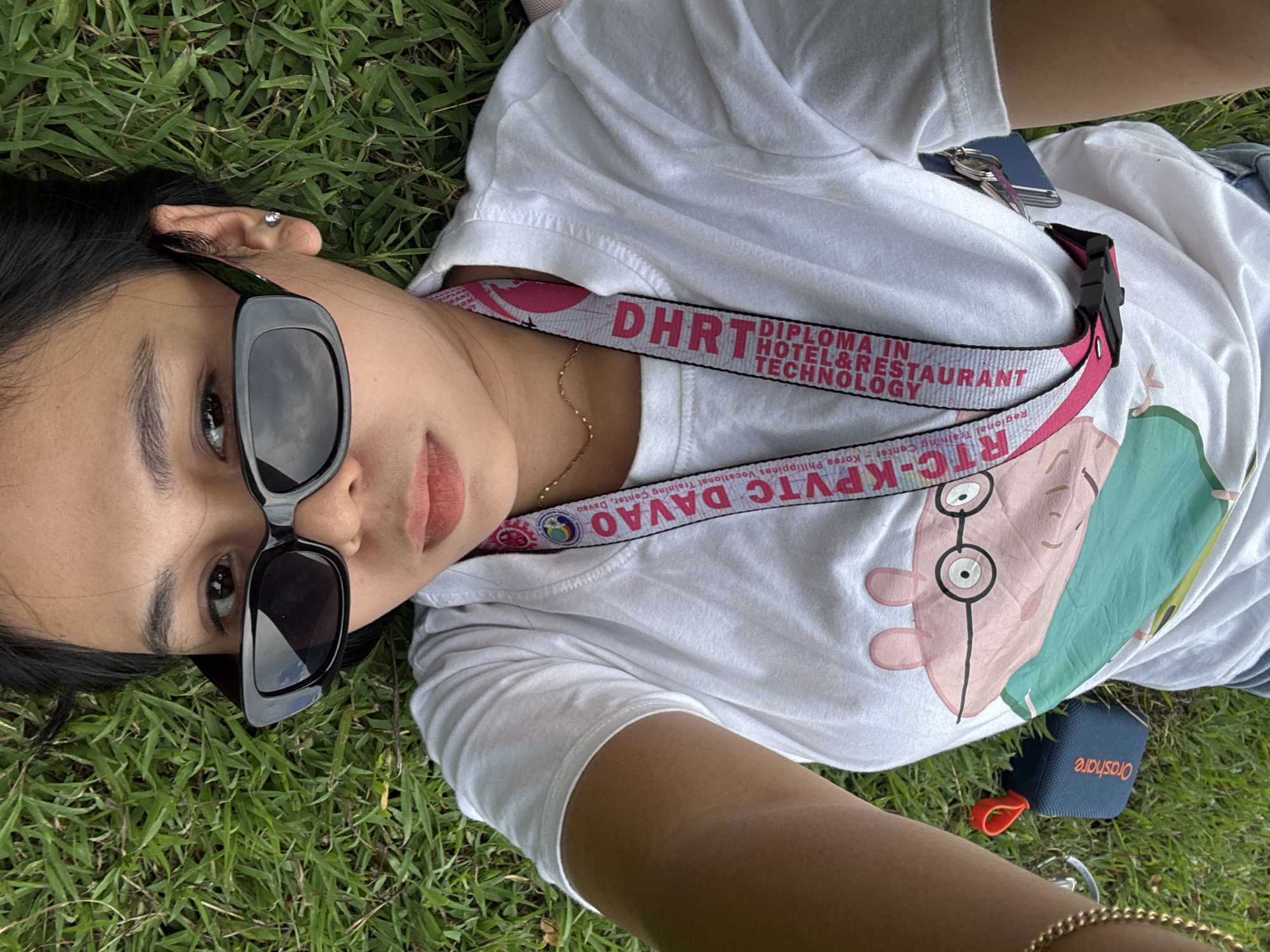 This is a close-up, high-angle shot of a person lying on green grass. The person, who appears to be young, is wearing a white t-shirt with a graphic print on it and black-framed sunglasses. A lanyard with "DH" and "RTC-KPVTC DAVAO" printed on it is visible around their neck, along with a small gold necklace. Keys and a blue lanyard are also tucked into the t-shirt.
The background is entirely composed of lush green grass. To the lower right of the frame, a small blue speaker with the word "Orashare" printed in orange is visible. The lighting suggests it could be daytime, possibly a bright day given the highlights on the sunglasses. The overall mood appears relaxed and casual. The visible text on the lanyard indicates a connection to the "RTC-KPVTC DAVAO - Regional Training Center - Korea Philippines Vocational Training Center, Davao," and "Diploma in Hotel & Restaurant Technology."