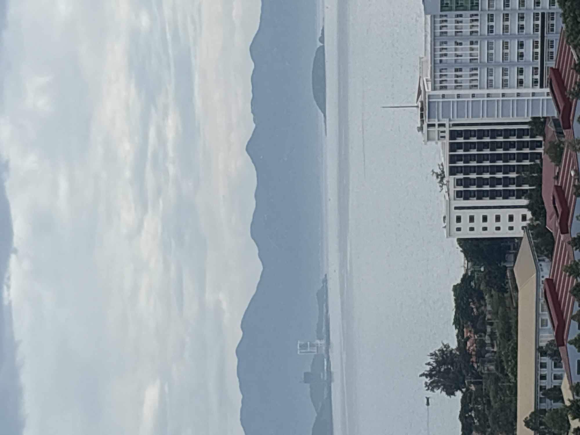 The image shows a view of the coastline of Nha Trang, Vietnam, with a body of water in the foreground, mountains in the background, and several buildings in the middle ground. The water appears calm, and there are a few small boats visible. The mountains are hazy, suggesting distance or a cloudy day. On the land, there are several modern buildings, including a tall white building with a distinctive geometric design and a cluster of smaller buildings with red roofs. The sky is overcast, with scattered clouds. The overall impression is of a coastal city with both natural beauty and urban development.