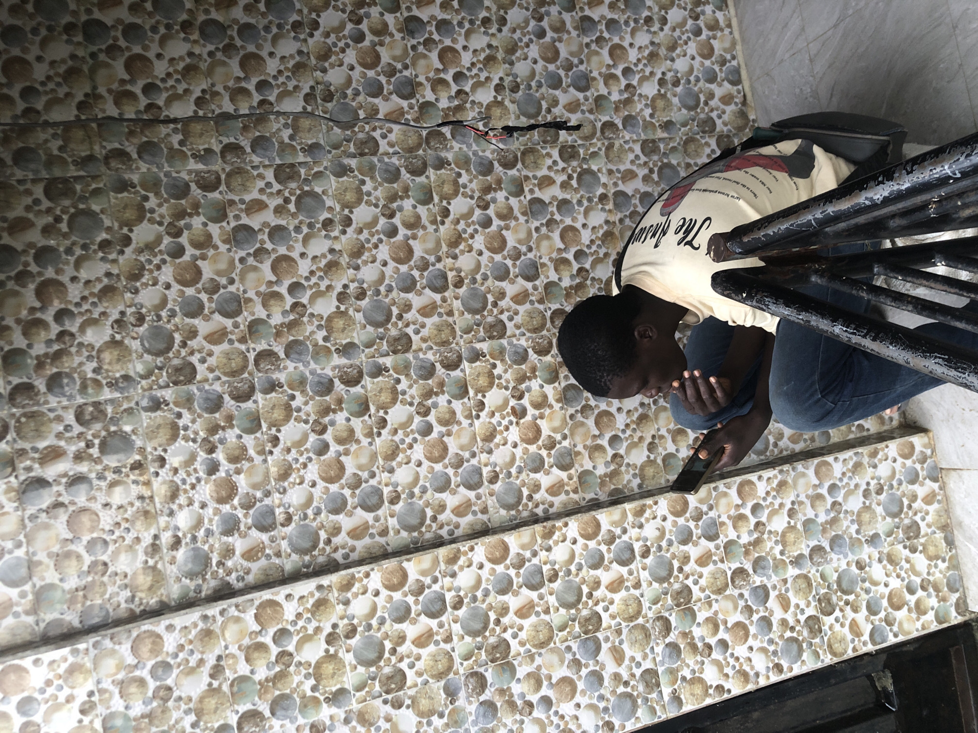 A young Black male is seated on concrete stairs, looking at a smartphone. He is wearing a light-colored long-sleeved shirt with text and graphics on the back, and blue jeans. The man is looking down at the phone, with his left hand supporting his chin and his right hand holding the phone. He is positioned to the right of the frame, with his body facing left. The background is a wall covered in mosaic tiles featuring circular patterns in shades of brown, beige, and gray. To the left of the man, a vertical tiled column is visible, and to the right, black metal railings are in the foreground. The scene appears to be outdoors, possibly on a porch or balcony, given the stairs and railing. The lighting suggests it is daytime. The overall environment is an urban setting, likely a residential or commercial area in Ado, Nigeria.
