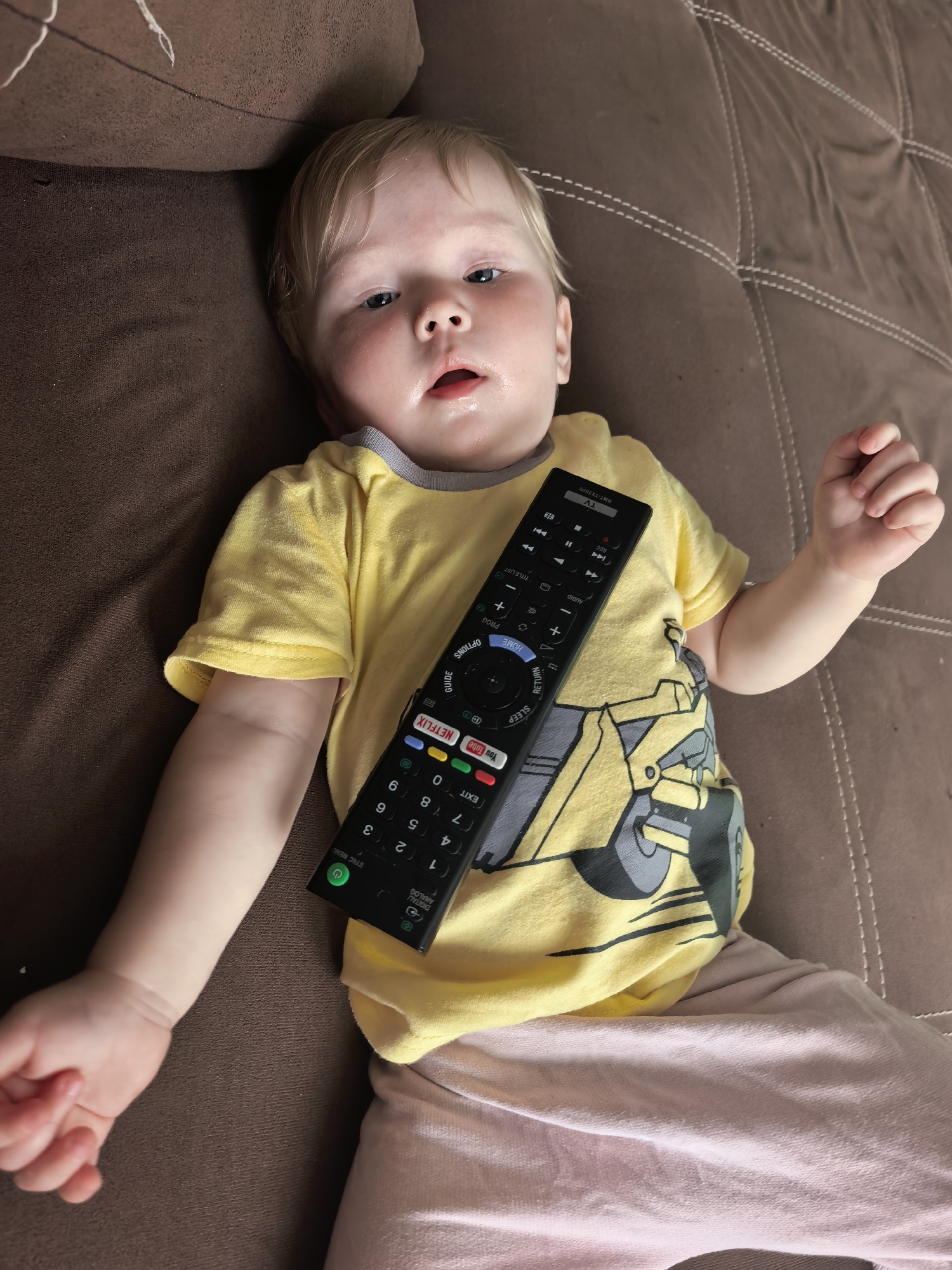 A baby wearing a yellow t-shirt with a yellow construction vehicle graphic and light pink pants lies on a brown textured surface, possibly a couch. The baby has blonde hair and appears to be looking upwards with a relaxed expression. A black television remote control rests diagonally across the baby's chest and torso. The background is a brown, tufted surface with visible stitching, suggesting an indoor setting. The image does not contain any discernible elements that would allow for the identification of its location within the city of Sterlitamak, Russia.