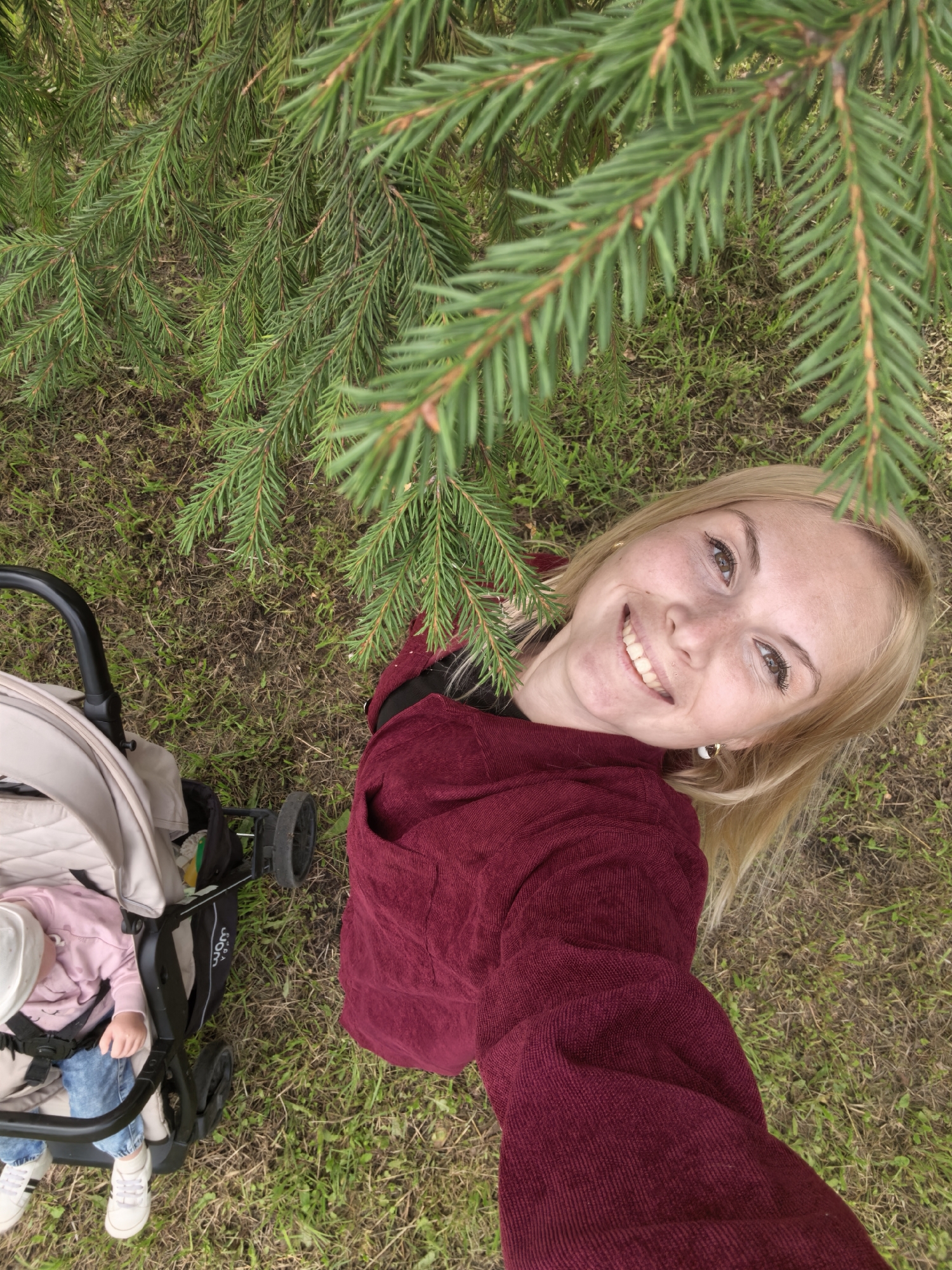 The image shows a top-down view of a woman and a baby in a stroller, outdoors. The woman, with blonde hair and freckles, is smiling and taking a selfie. She is wearing a maroon sweater. Next to her is a baby in a beige stroller, wearing a pink shirt, blue jeans, and a white hat. The stroller has black wheels and a black handle. The background is a grassy area with scattered dry leaves and pine needles, indicating a park or wooded area. The bright green needles of a pine tree hang over the scene from the top of the frame. The overall lighting suggests daylight. The location is unspecified but is in the city of Sterlitamak, Russia.