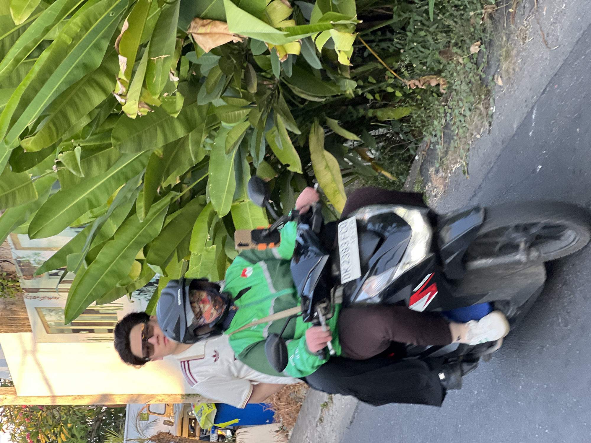 Two individuals are riding a motorcycle on a paved road in North Kuta, Indonesia. The person in the front, wearing a green jacket and a helmet with a patterned mask, is driving the motorcycle. The person in the back, wearing a white Adidas-branded shirt and sunglasses, is a passenger. They are surrounded by lush green foliage, suggesting a tropical environment. The time of day appears to be late afternoon, indicated by the warm sunlight casting shadows. The license plate on the motorcycle is visible, showing "DK 2383 FCG".