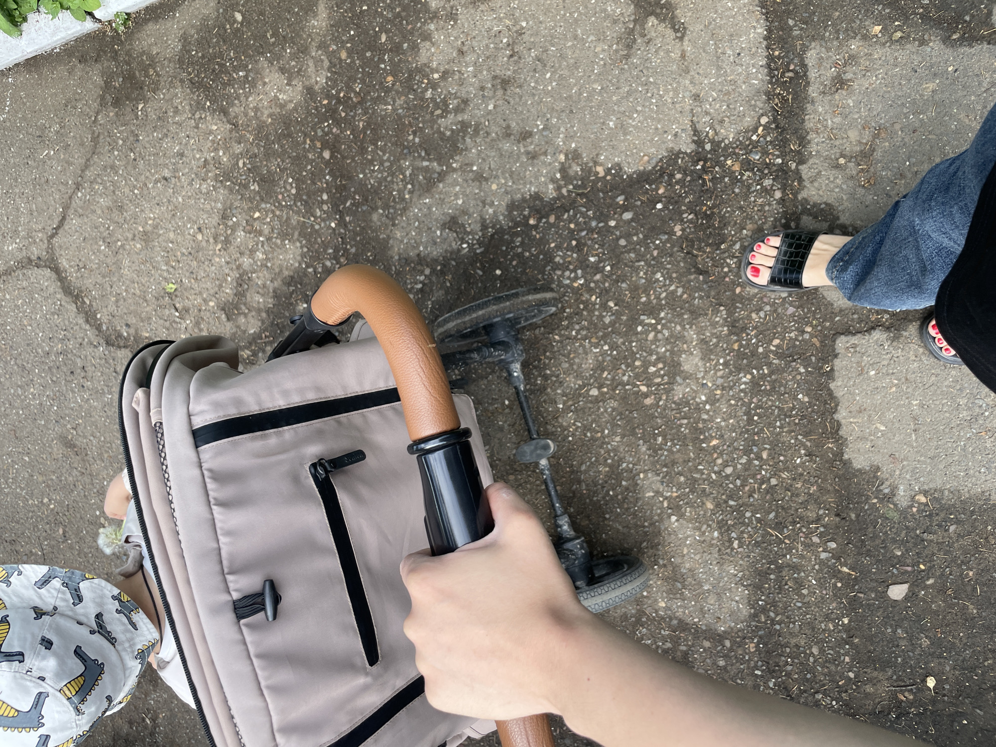 The image is taken from a first-person perspective, looking down at a concrete sidewalk in an urban environment. The person taking the photo is holding the handle of a beige baby stroller. A child wearing a dinosaur-patterned bucket hat is partially visible in the stroller. To the right, there is a woman's foot wearing a black sandal with red nail polish, and she is wearing blue jeans. The sidewalk is mottled and has some visible cracks and debris. The lighting suggests it is daytime.