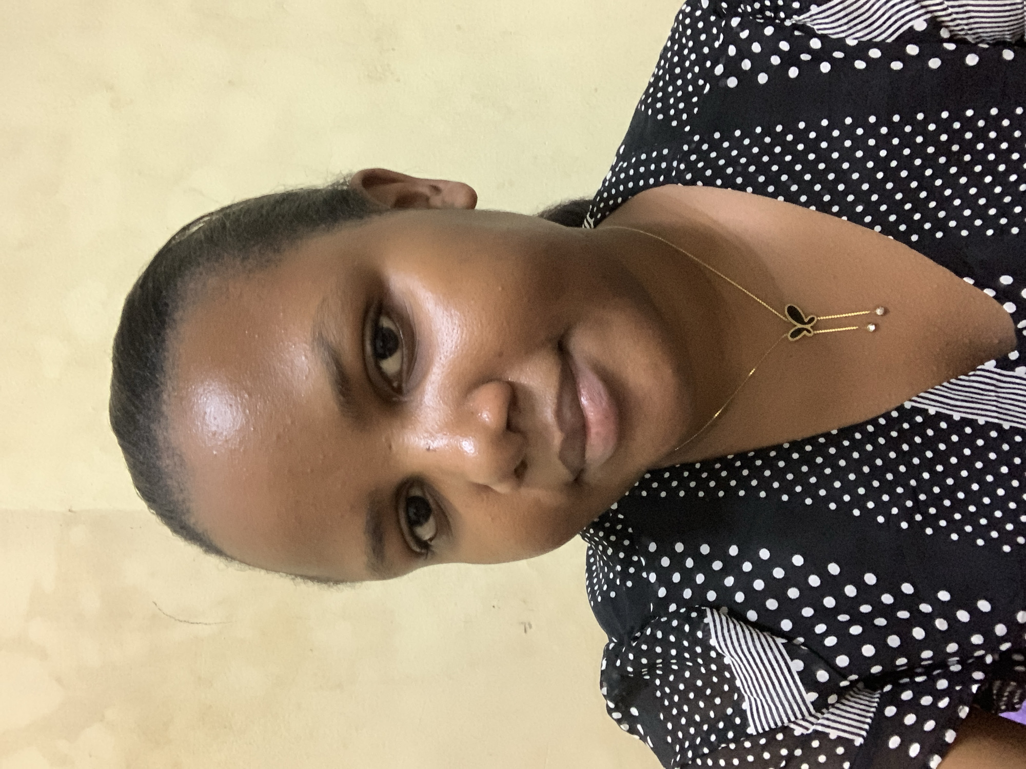 A medium close-up shot depicts a woman with dark skin. She is facing slightly toward the right side of the frame, and she gazes directly at the camera with a gentle smile. Her hair is pulled back, and she wears a gold necklace featuring a butterfly pendant. Her attire consists of a black and white polka-dot top with striped accents.
The background is a plain yellow wall. There is no information to suggest that this photo was taken in Dar es-Salaam, Tanzania.