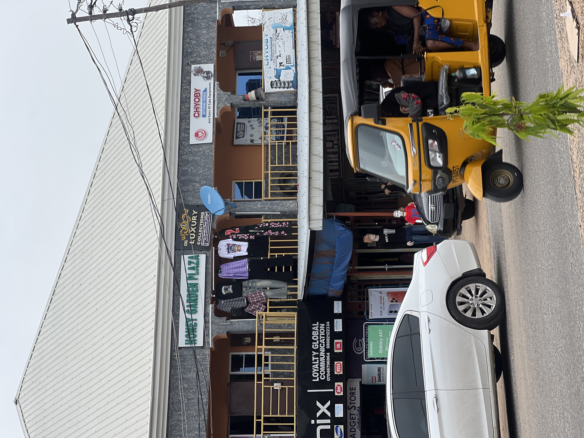 A street scene in Jalingo, Nigeria, features a multi-story building with businesses on the ground and upper floors. The building has a corrugated metal roof. A white sedan is parked on the left, and a yellow auto-rickshaw with passengers is in the foreground. A black car is partially visible behind the auto-rickshaw.
Signs on the building advertise "HONEY GARDEN PLAZA," "DE LUXURY COLLECTIONS," and "CHYOBY CATERING SCHOOL And Sales Of Cake Equipments." Below these, a banner for "LOYALTY GLOBAL COMMUNICATION" is displayed, listing phone numbers and various mobile brands. Further down, there are advertisements for Samsung Galaxy A07 and Tecno POP 20 SPARK 50 phones. Mannequins are visible in front of some shops, one wearing a red shirt with the number "29" and another in a black long-sleeved shirt with the word "REPLY".
The street is paved, and power lines are strung across the scene. Vegetation, including a small leafy plant, is present on the side of the road. The sky is overcast.