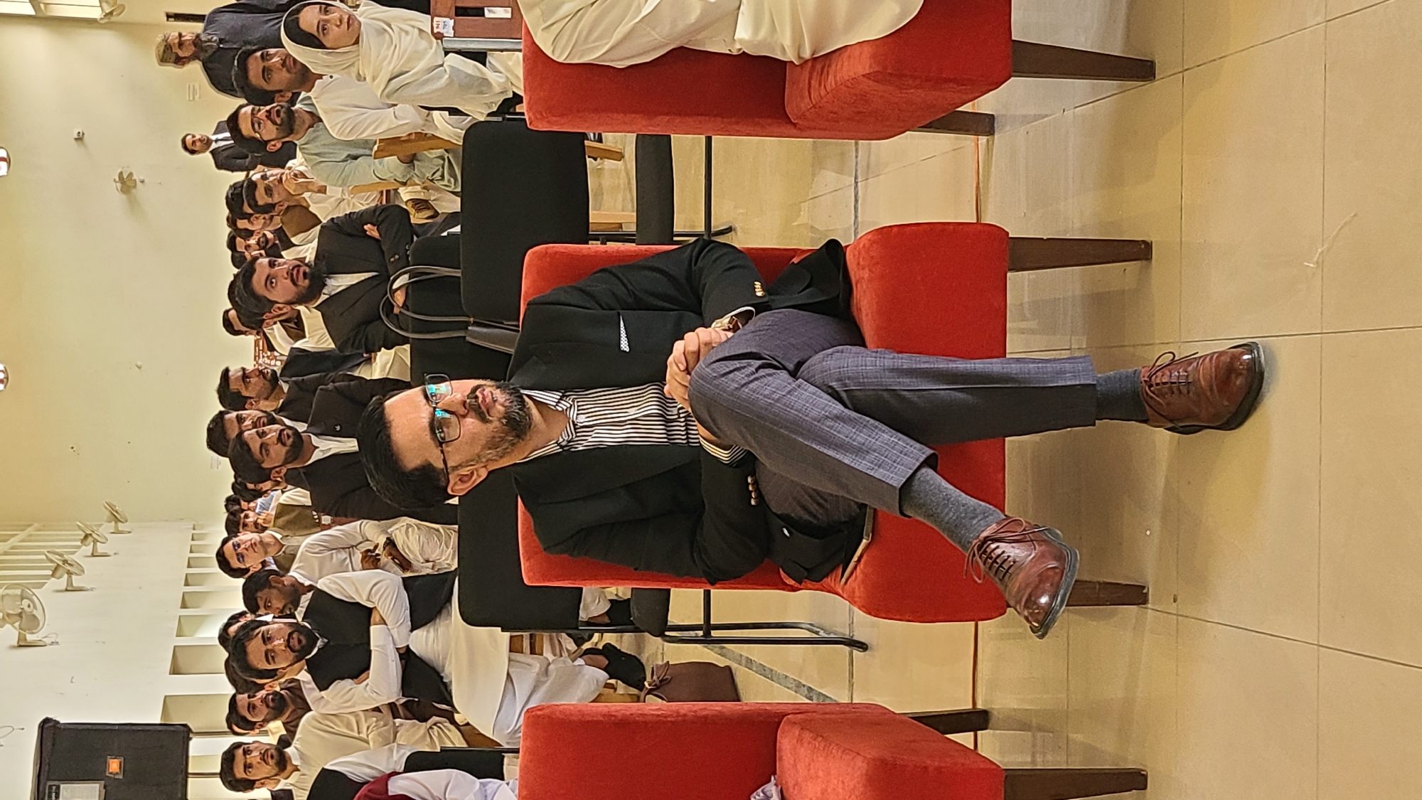 The image depicts a gathering of people in Quetta City Tehsil, Pakistan. The setting appears to be an auditorium or a large room with tiled floors and a row of wall-mounted fans visible in the background. A man in a dark suit, striped shirt, glasses, and brown shoes is reclining on a red chair with his legs crossed. He is the focal point of the image. Behind him, rows of men are seated, many dressed in dark suits or traditional Pakistani clothing. At the top of the frame, a woman is seated, wearing a white headscarf. The overall impression is of a formal or semi-formal event taking place in the city.