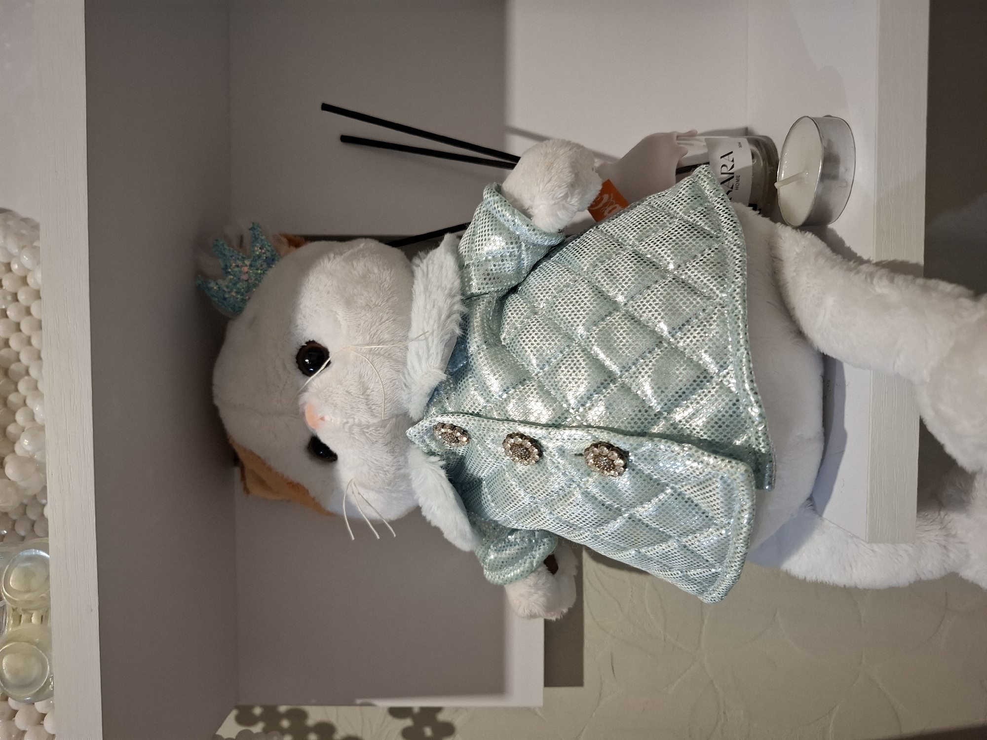 A plush white and orange cat toy wearing a shimmering blue jacket with three decorative buttons sits on a white shelf. The cat has large black eyes, a pink nose, and white whiskers. It also wears a small, light blue crown with glitter on its head. Behind the cat, on the same shelf, are several thin black sticks protruding from a bottle, a small white tea light candle in a metal holder, and a clear bottle with an orange tag. Above the shelf, out of focus, are several clear glass containers filled with small white orbs. The background wall to the left has a textured pattern. The image was taken in Korenovsk, Russia.