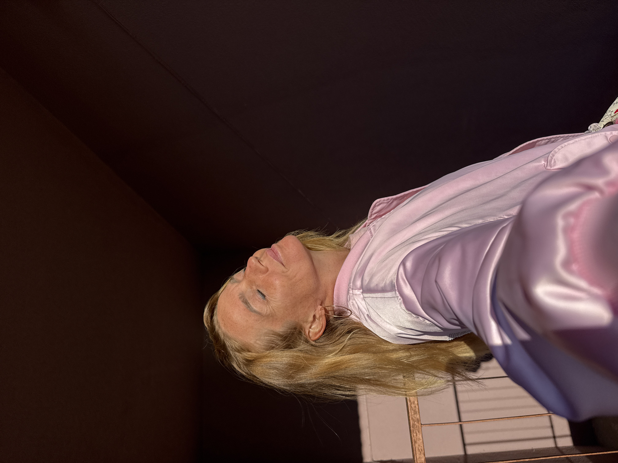 A blonde woman stands with her eyes closed, facing slightly upwards and to the right, as if basking in sunlight. She is wearing a shimmering pink bomber jacket. She has hoop earrings on. Behind her, a dark wall curves away, and to her left, a metal railing casts a shadow on a light-colored wall. The image appears to be taken outdoors, possibly in a courtyard or on a balcony in Sinyavino, Russia.