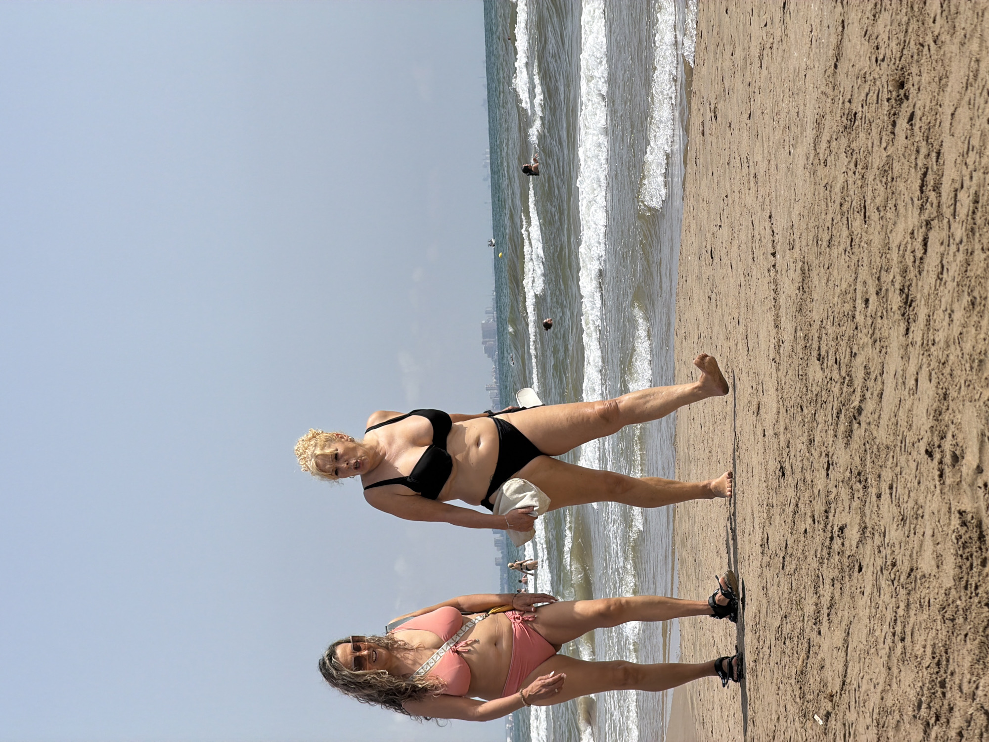 Two adult women are walking on a sandy beach. The woman on the left is wearing a pink bikini and sunglasses, and has curly brown hair. The woman on the right is wearing a black bikini and has blonde curly hair. She is holding a light-colored towel and a white object. The ocean with waves is visible in the background, and there are other people in the water. In the distance, a city skyline is visible on the horizon under a clear sky.