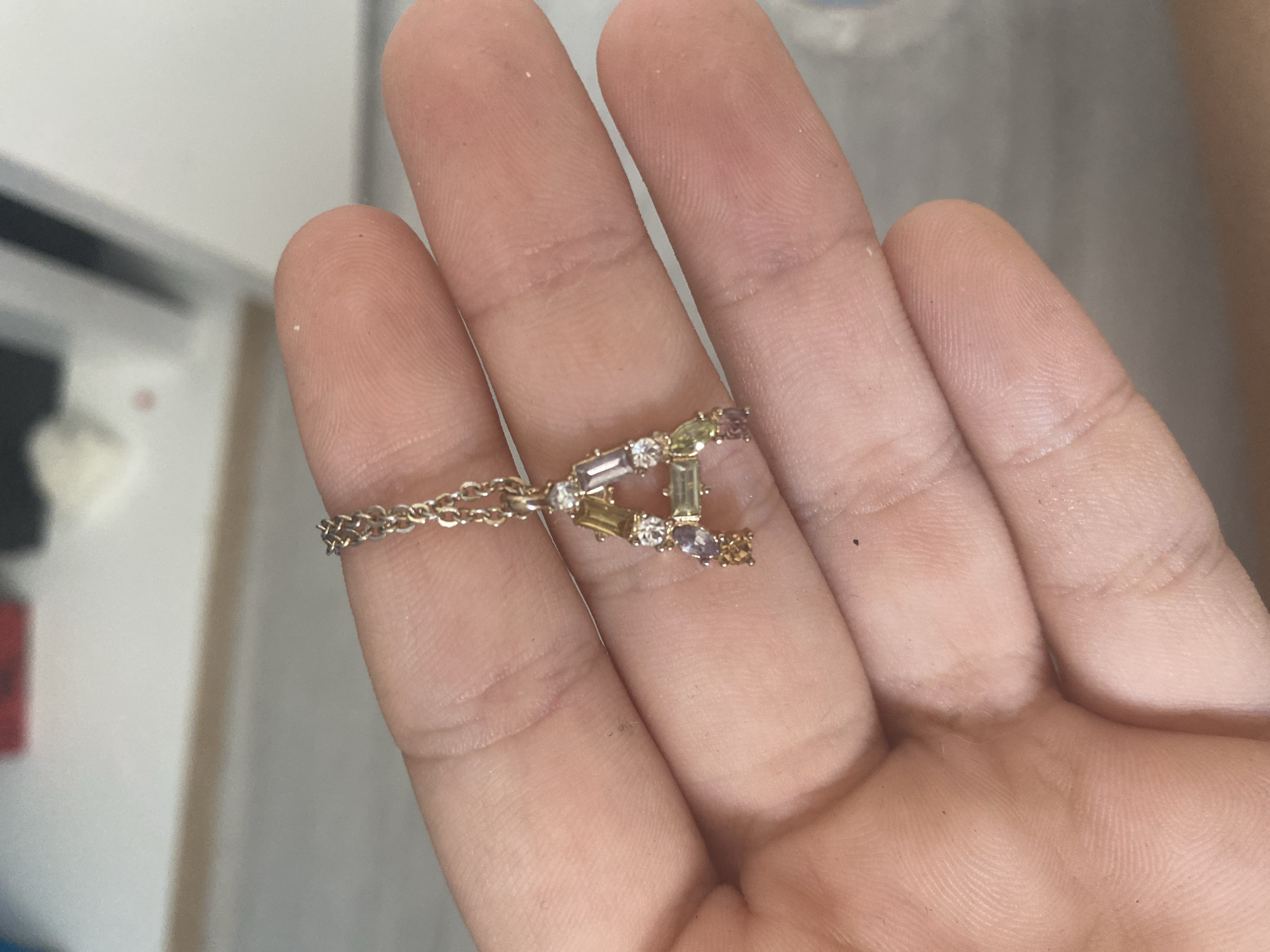 A close-up, top-down shot shows a person's hand, which is slightly aged and has visible fingerprints, holding a delicate gold ring. The ring is adorned with small, baguette-cut gemstones in various colors, including purple, light green, clear, and yellow, arranged in an abstract, geometric pattern. A fine gold chain extends from the ring, creating a bracelet-like effect around the wearer's finger. The background is a soft blur of light gray and white, suggesting an indoor setting. No specific location in Blagoevgrad, Bulgaria, is identifiable from the image.