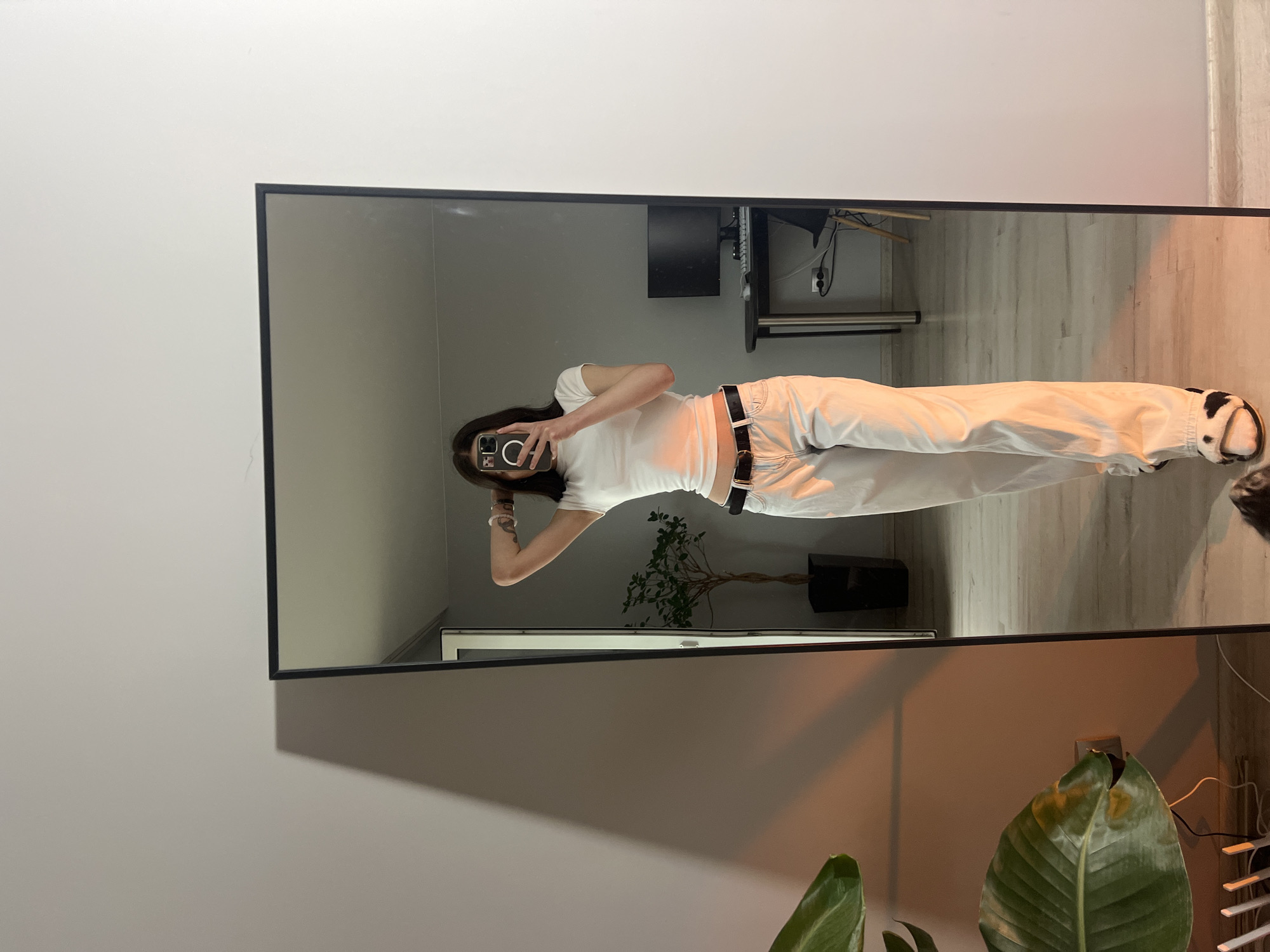 A young person is captured in a full-length mirror selfie within an indoor setting in Ternopil, Ukraine. The person, with long dark hair, is reflected standing upright, wearing a plain white short-sleeved t-shirt, light-colored wide-leg pants, and a black belt. They are holding a smartphone to take the picture, with part of their face obscured by the phone. A tattoo is visible on their right upper arm. On their left foot, they wear a black and white animal-print slipper or shoe.
The mirror has a slim black frame and reflects a modern room. In the background, visible both in the reflection and directly, is a light-colored wooden floor and neutral-toned walls. Reflected behind the person is a desk with a computer monitor, cables, and what appears to be a power strip. A small potted plant with sparse branches is also reflected. To the bottom right of the mirror, a larger plant with broad green leaves, some showing browning, is directly visible. A warm, orange-toned light source illuminates the right side of the image, casting a glow on the floor and walls. The overall ambiance is casual and tidy, indicative of a residential space. There is no visible text in the image.