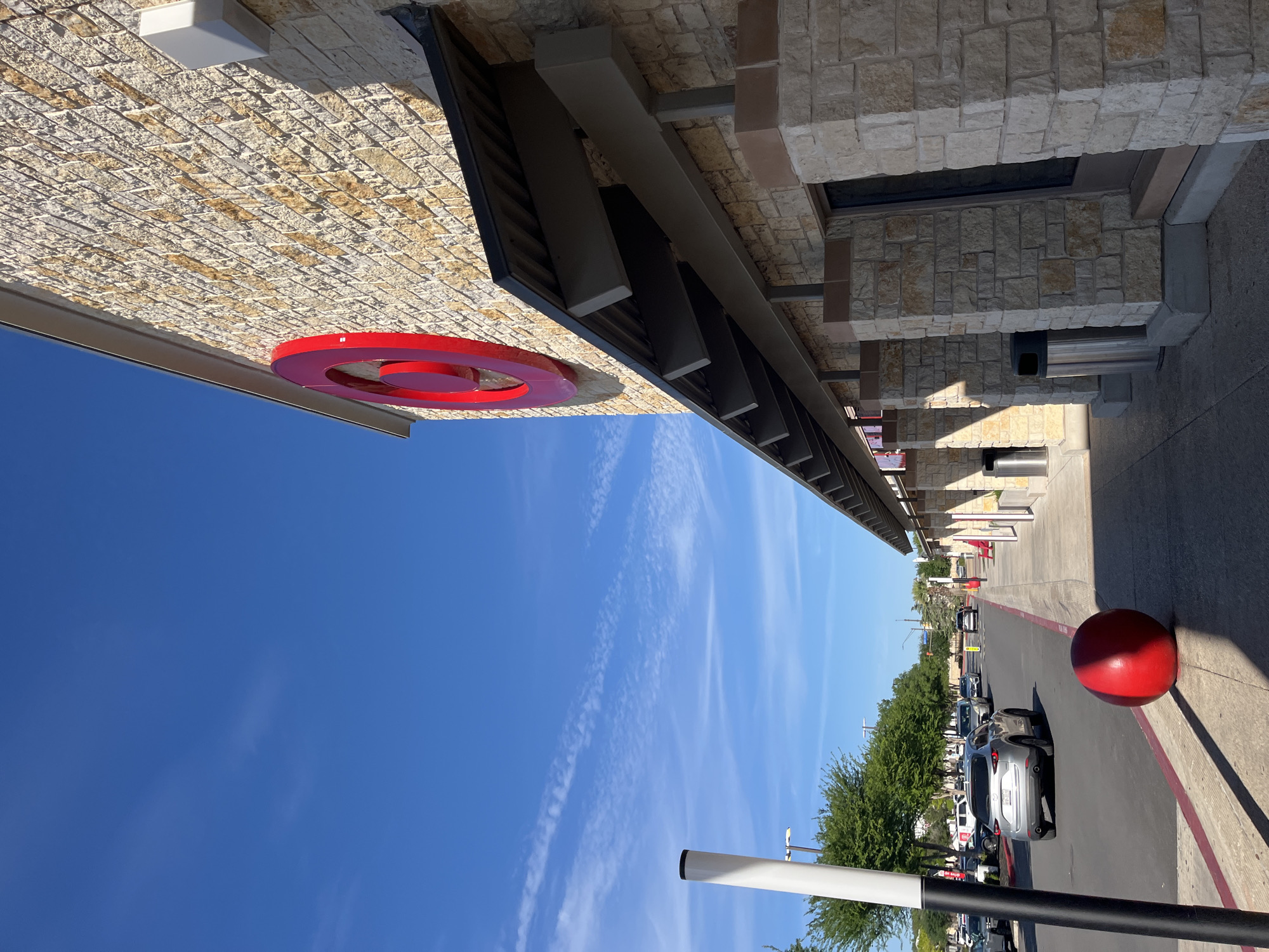 The image depicts the exterior of a Target store in San Antonio, United States. The building is constructed with tan and gray stone and has a red Target logo near the roofline. A red decorative sphere is visible near the entrance, adjacent to parked cars. The sky is bright blue with wispy clouds.