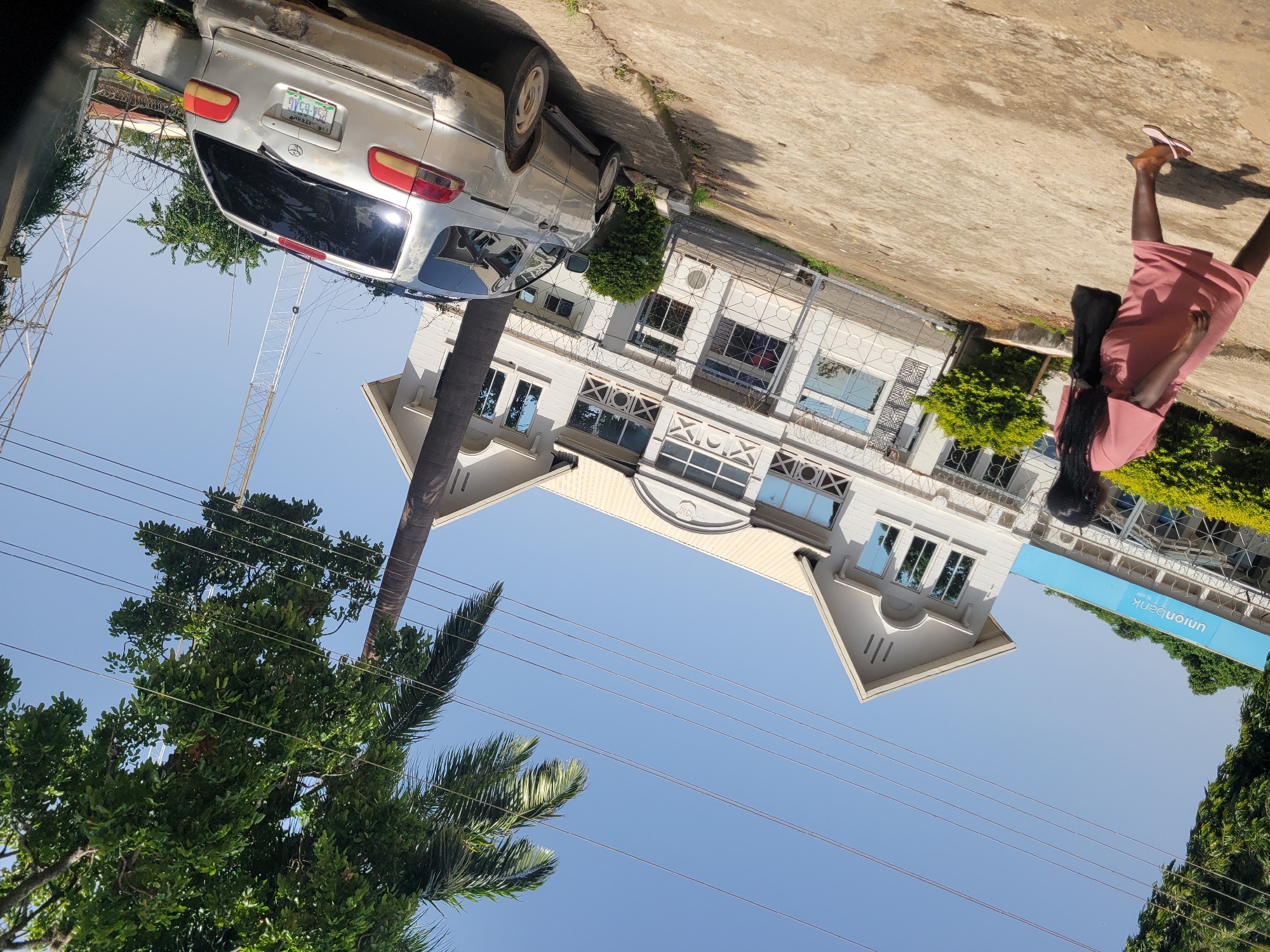 This is a rotated, eye-level, outdoor, daytime photograph taken in Jos, Nigeria, showing a street scene. In the foreground, a woman wearing a pink dress and pink sandals walks away from the camera, carrying a black bag over her left shoulder. To her left, a light blue sign with white lettering that reads "unionbank" is visible. Behind the woman, a modern, multi-story building with white walls and numerous windows is depicted. Adjacent to the building and partially obscuring it is a silver minivan. The vehicle has a visible license plate. Overhead, several power lines stretch across a clear blue sky. Tall trees, including a palm tree, are also present, with their branches reaching into the frame from the left side. In the background, industrial-looking structures, possibly telecommunication towers, can be seen against the sky. The ground is a paved surface with some cracks and discoloration.