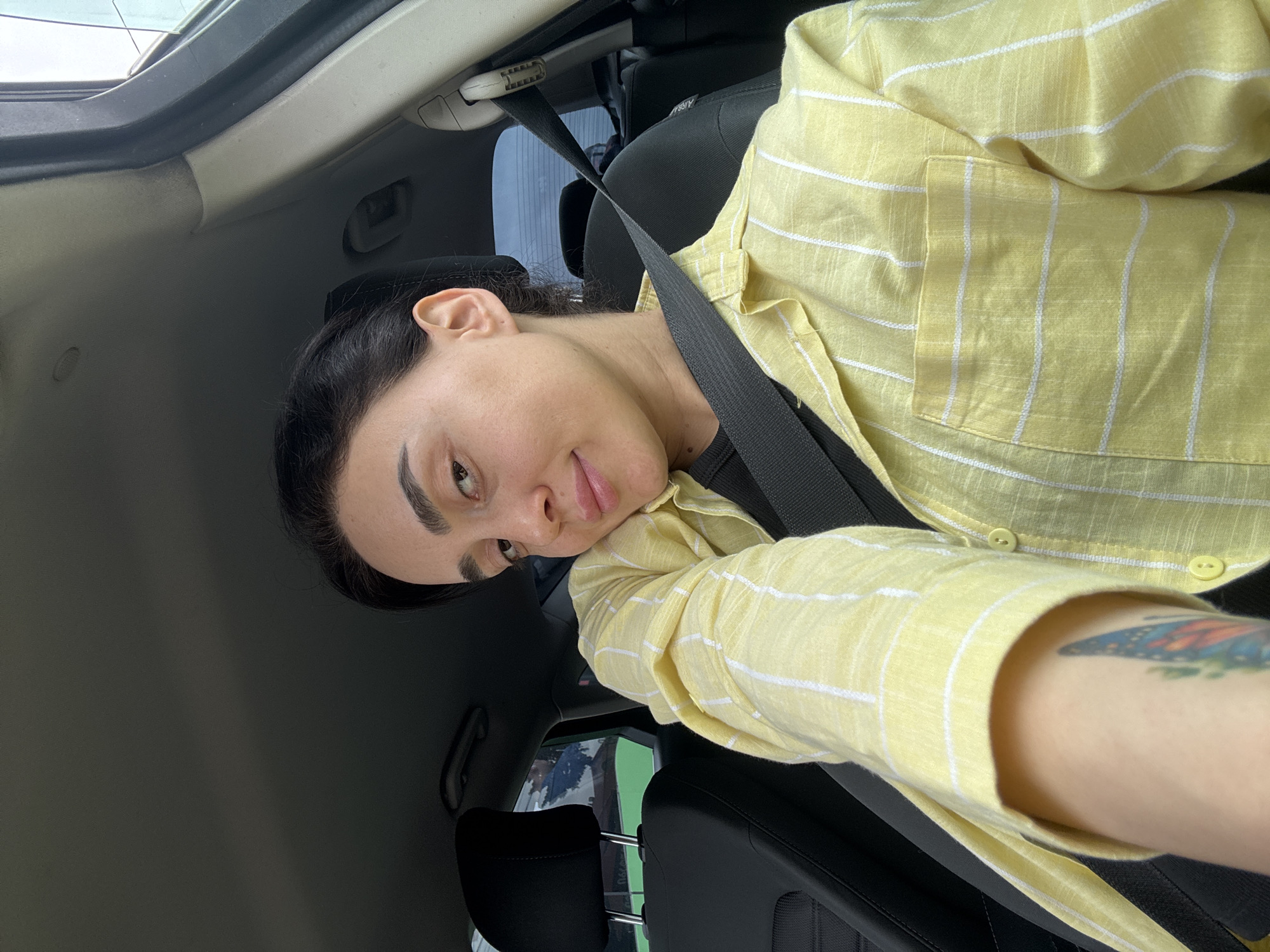 A young adult is captured in a tilted selfie, seated in the passenger side of a car in Tbilisi, Georgia. The person has dark hair, styled away from their face, and a soft, closed-mouth smile, conveying a relaxed expression. They are wearing a light yellow button-up shirt with thin white vertical stripes over a dark undershirt. A black seatbelt crosses their chest and shoulder, indicating they are buckled in.
The car's interior features a black seat with the text "AIRBAG" visible on its side, and a grey headliner and pillar. Through the car window, glimpses of outdoor greenery or a building can be seen, indicating it is daytime. A colorful tattoo, resembling a butterfly wing, is visible on their left forearm, peeking out from the sleeve of their shirt. The overall impression is a casual, candid moment during a car journey.