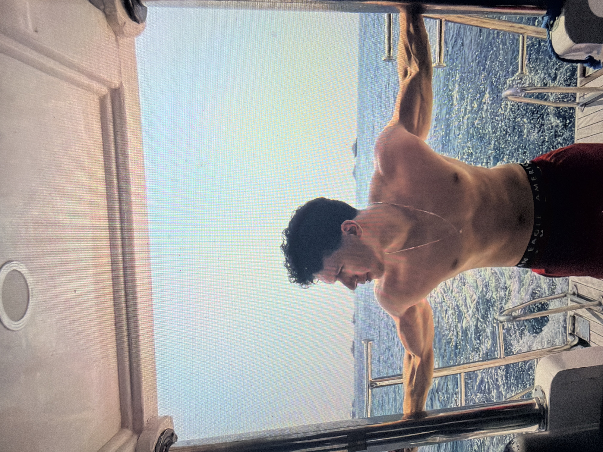 This image depicts a young, shirtless male with dark, curly hair and a muscular physique, standing on the stern deck of a boat. He is positioned in profile, looking towards the right side of the frame, with both arms extended horizontally, gripping vertical metal stanchions that flank the opening of the boat's rear. He wears red shorts with the "AMERICAN EAGLE" brand visible on the waistband, and a thin chain necklace is draped around his neck. The boat's deck appears to be made of light-toned wooden or composite slats, with integrated metal steps and additional railings leading down into the water. Behind the boat, the sea is visibly choppy with whitecaps, indicating motion, under a bright, clear light-blue sky. Several distant vessels are discernible on the horizon line. The overhead structure of the boat's canopy or cabin ceiling, colored white with decorative paneling and a circular recessed light fixture, is visible in the upper portion of the frame. The entire image displays a prominent pixel grid pattern, characteristic of a photograph taken of a digital screen. The location is in the city of Shumen, Bulgaria.
