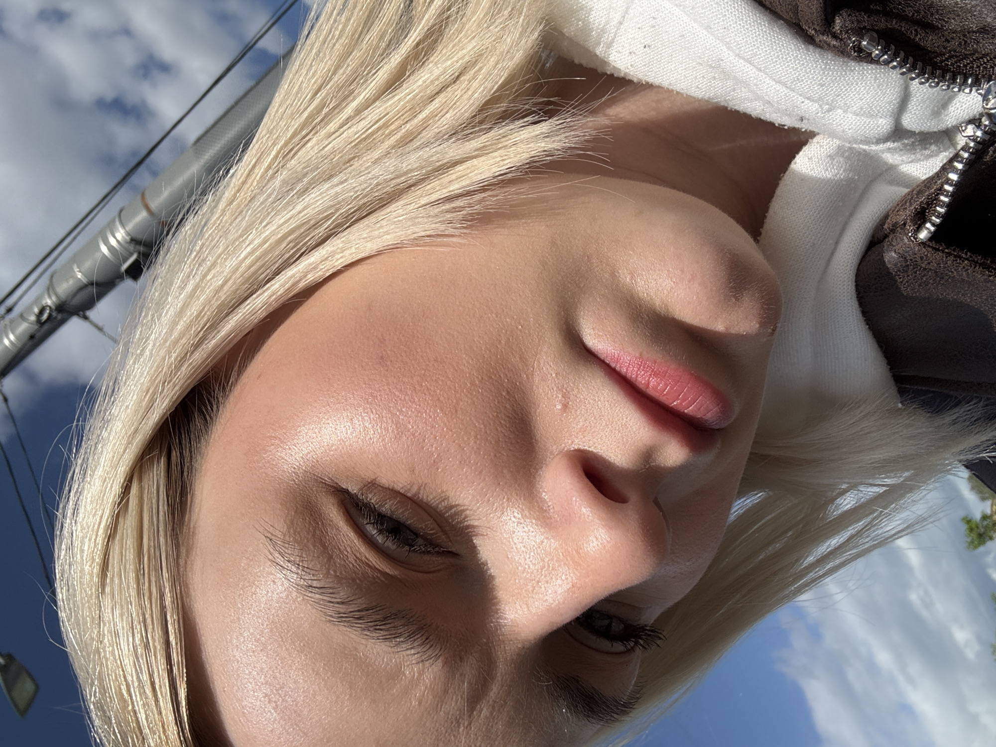 The image shows a close-up of a person with light blonde hair, fair skin, and light pink lips. They are wearing what appears to be a white and brown zip-up jacket. The background features a blue sky with scattered clouds, and a utility pole with wires is visible. The sky and pole suggest an outdoor setting, possibly urban. Without more contextual clues, I cannot determine if the image was taken in Saint Petersburg, Russia.