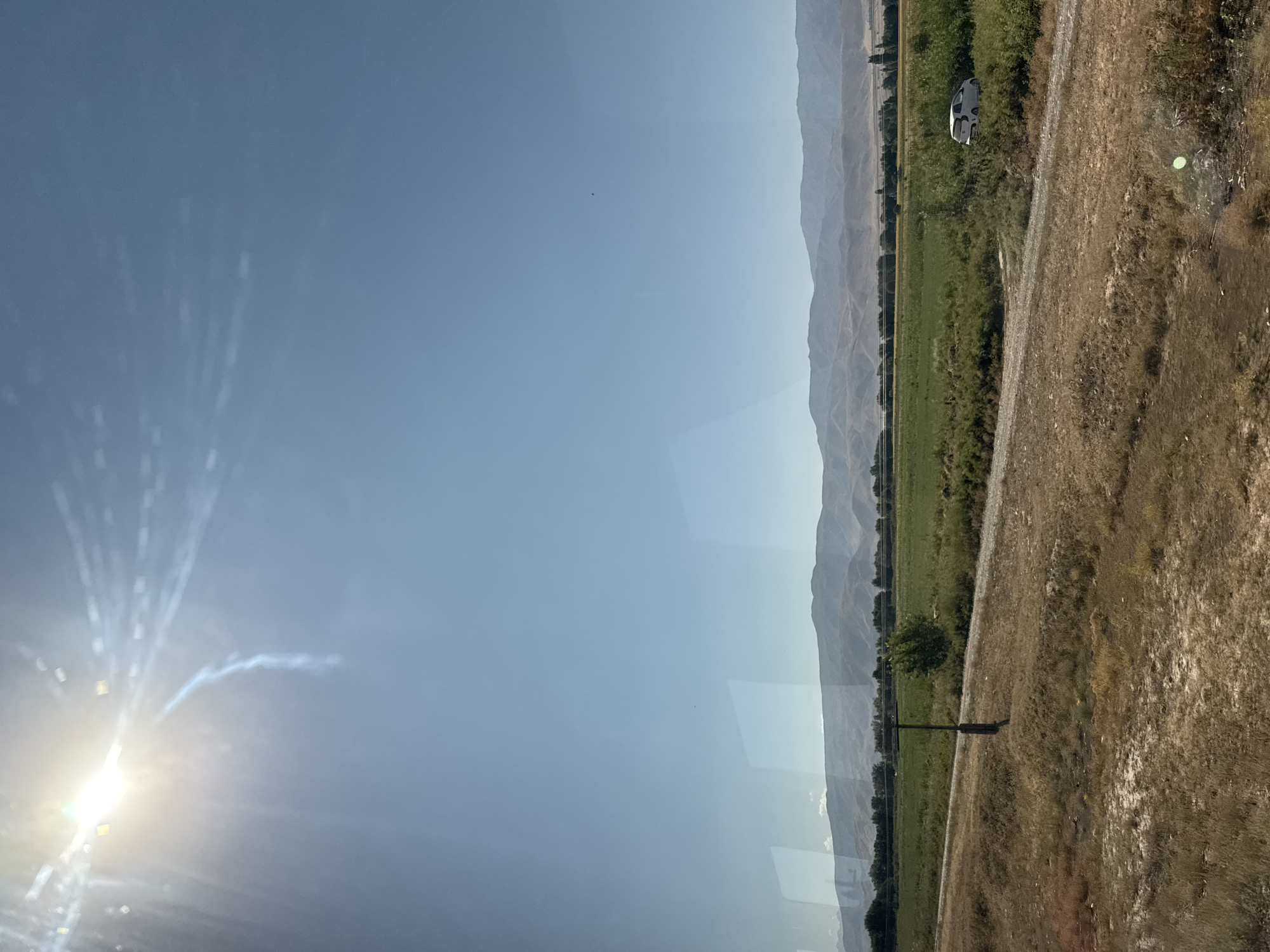A bright sun shines in the upper left corner of the image, casting a lens flare across the clear blue sky. Below the sky, a range of mountains stretches across the horizon. In the midground, a field of green grass is bordered by a dirt road. A lone telephone pole stands in the field, and a white car is parked in the brush to the right. The foreground is a dry, grassy plain. The scene is reminiscent of the rural outskirts of Orlovka, Kyrgyzstan, during a clear, sunny day.