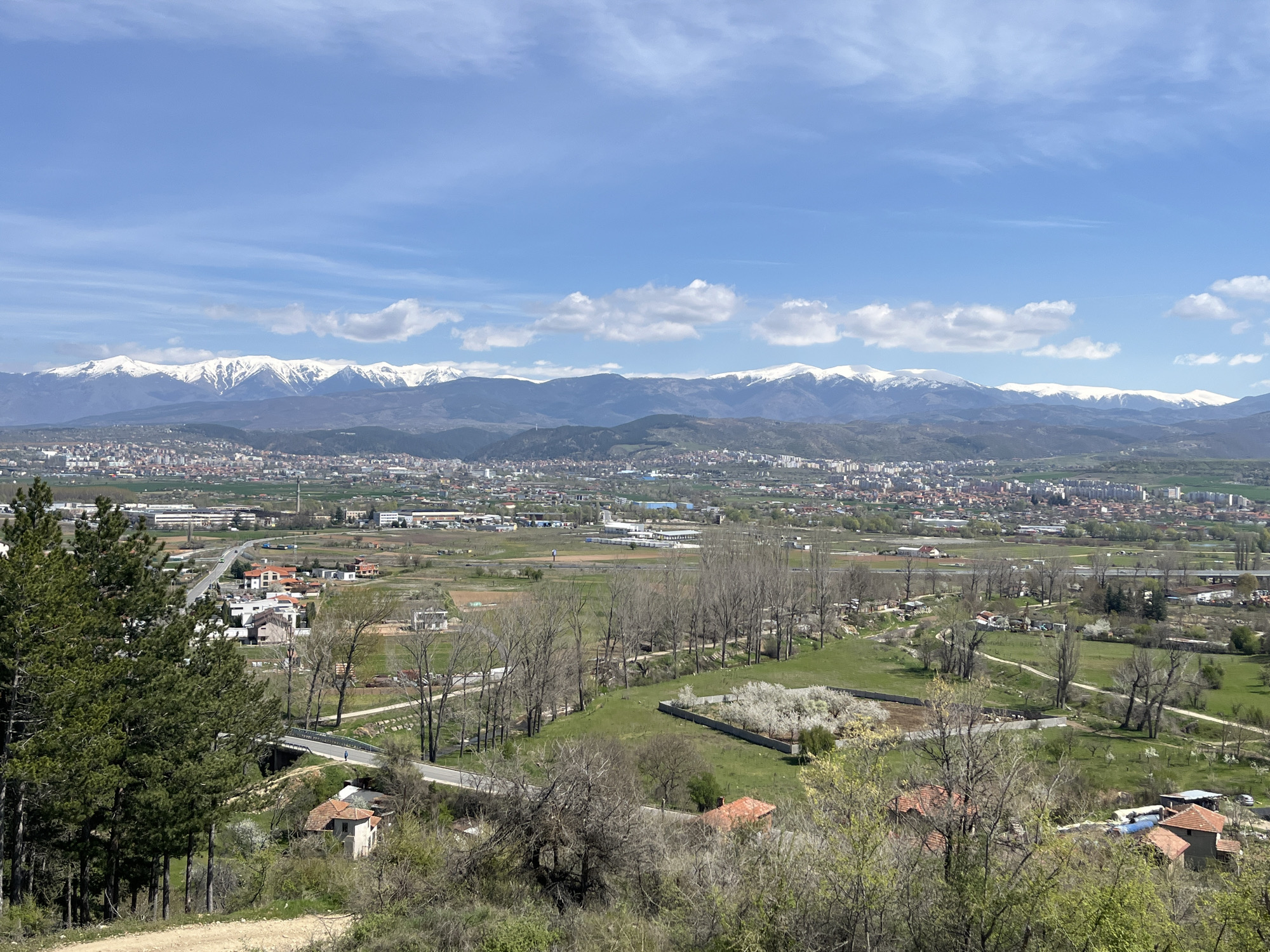 The image shows a panoramic view of a city nestled in a valley, with snow-capped mountains in the distance. The city appears to be sprawling, with a mix of residential and industrial areas. In the foreground, there is a more rural landscape with green fields, trees, and scattered houses. A road and a bridge are visible in the lower left. The sky is blue with scattered white clouds. The exact location and city are not identifiable from the image alone.