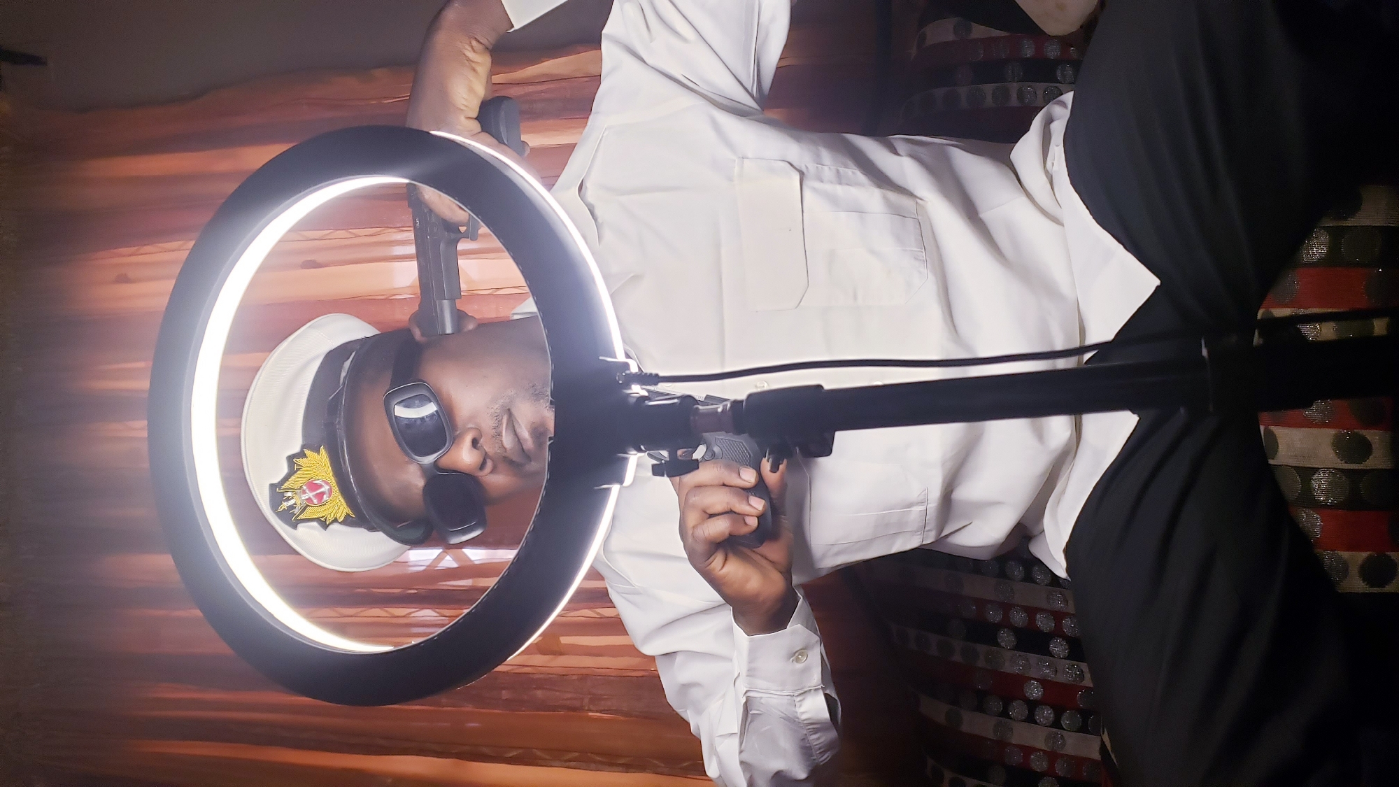 An indoor, portrait-style shot, rotated to appear sideways, features a man of African descent posing in front of a brightly illuminated, circular ring light. He is dressed in a crisp, long-sleeved white shirt and dark trousers. On his head, he wears a white peaked cap adorned with a detailed gold and red emblem, suggesting a military or naval uniform. His eyes are obscured by dark sunglasses, adding to a serious or composed demeanor.
The man's hands are actively engaged with the equipment. His left hand holds a smartphone, likely mounted on a grip or stand attached to the ring light, positioned to record. His right hand grasps another black device, possibly a remote control or part of the light setup. A black microphone on a slender boom arm extends from the ring light assembly towards his face, indicating an audio recording or live streaming setup.
The background consists of patterned fabric, likely drapes or a backdrop, featuring horizontal stripes and circular motifs in shades of red, black, and silver/grey. The ring light provides the primary and most prominent illumination for the scene, creating a soft, even light on the subject. The overall impression is that of an individual engaged in media creation, such as vlogging, live streaming, or a professional photoshoot. No specific location cues are visible within the image itself, though the context provided states the location is Sabon Tasha, Nigeria. No visible text is clearly legible on any object.