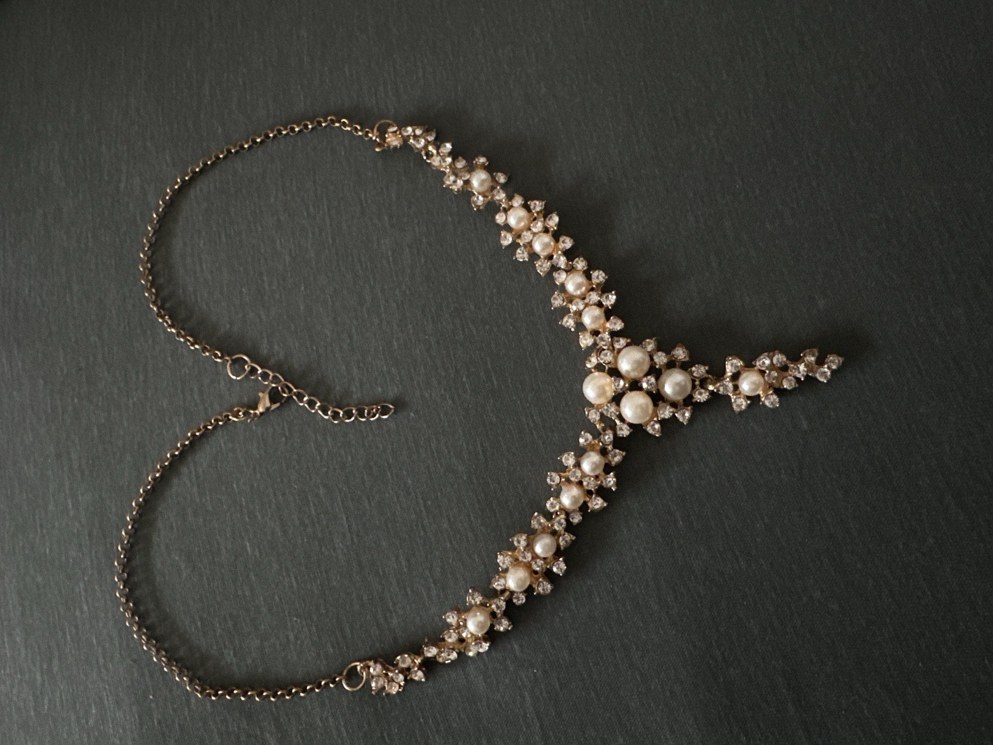 The image features an elegant necklace displayed flat against a dark, textured grey background, possibly a stone surface. The primary subject is a gold-toned necklace adorned with pearls and clear gemstones.
The necklace has a delicate chain on both ends, with an adjustable clasp and extender chain visible on the left side. The central and most prominent part of the necklace is intricately designed, featuring numerous small, round, white or cream-colored pearls. Each pearl is set within a cluster of sparkling clear rhinestones or crystals, forming a series of symmetrical, flower-like or star-like motifs. These ornate segments are connected along the length of the necklace, creating a bib-style or collar design that widens towards the center.
No specific activity is taking place; the necklace is presented as a static object, likely for product photography or personal admiration. The lighting is soft and even, highlighting the luster of the pearls and the sparkle of the gemstones without casting harsh shadows. No text is visible within the image. This media file is associated with the location context of Riga, Latvia.