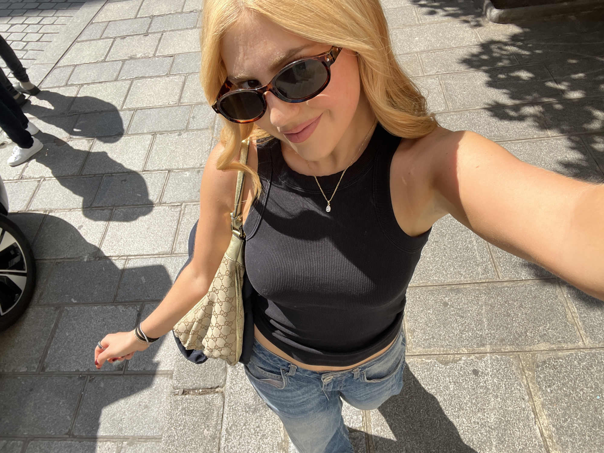 A woman stands on a gray tiled street, taking a selfie. She is wearing sunglasses, a black tank top, and blue jeans. A light-colored Gucci handbag hangs from her left shoulder. She also wears a gold necklace and multiple bracelets on her left wrist. A shadow from a tree or building is cast to the right of her, and the legs and shoes of another person are visible on the left side of the frame, alongside a car tire. The location is likely in a city based on the paved street and building shadows, but more information is needed to specifically identify it as Paris.