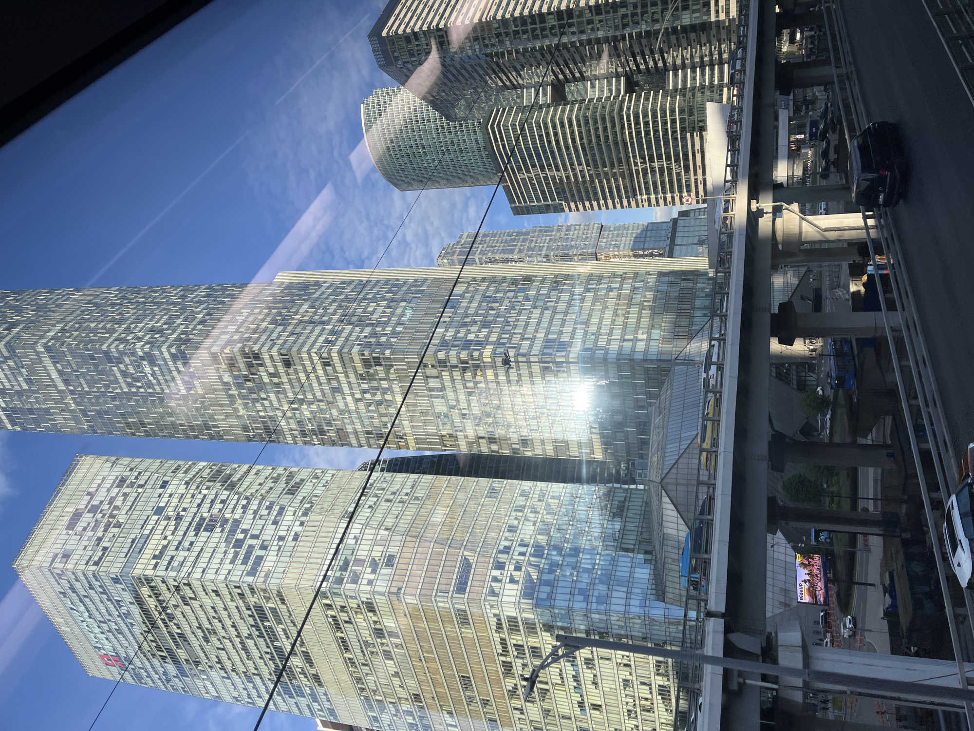 This image shows a view of modern skyscrapers in Moscow, Russia, from a high vantage point, possibly from a vehicle or elevated walkway. The buildings are characterized by their glass facades, reflecting the blue sky and sunlight. Several distinct architectural styles are visible, including a curved glass skyscraper and a rectangular tower with a pattern of horizontal and vertical lines on its exterior.
The scene is bathed in bright sunlight, suggesting it is daytime, likely mid-morning or afternoon, with a clear blue sky and some faint contrails from an aircraft. A multi-level road or elevated highway runs through the scene, with cars and a black SUV visible on it. The perspective is slightly tilted, giving a dynamic feel to the urban landscape.
In the distance, more buildings and infrastructure can be seen, indicating a dense urban environment. One of the building's facades features a digital billboard displaying an advertisement with text that includes "PICK-UP" and "PIZZA". The overall impression is one of a bustling, modern metropolis. The reflection on the glass surfaces creates a sense of depth and complexity, blending the architectural structures with the sky and surrounding environment.