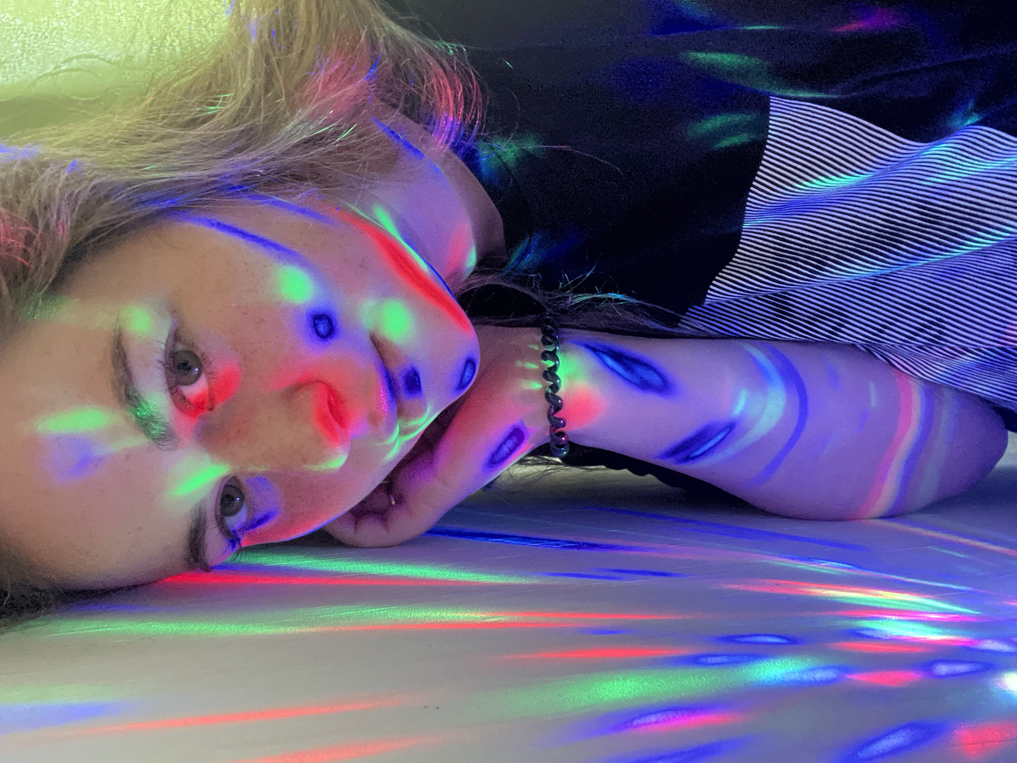 A young person with blonde hair is lying on a white surface, illuminated by multicolored lights. The lights are casting colorful streaks and patterns across their face, hand, and the surface they are lying on. The person is wearing a dark shirt and a striped sleeve or garment is visible. A bracelet is on their wrist. The location in the city of Сосново, Russia is not apparent from the image.