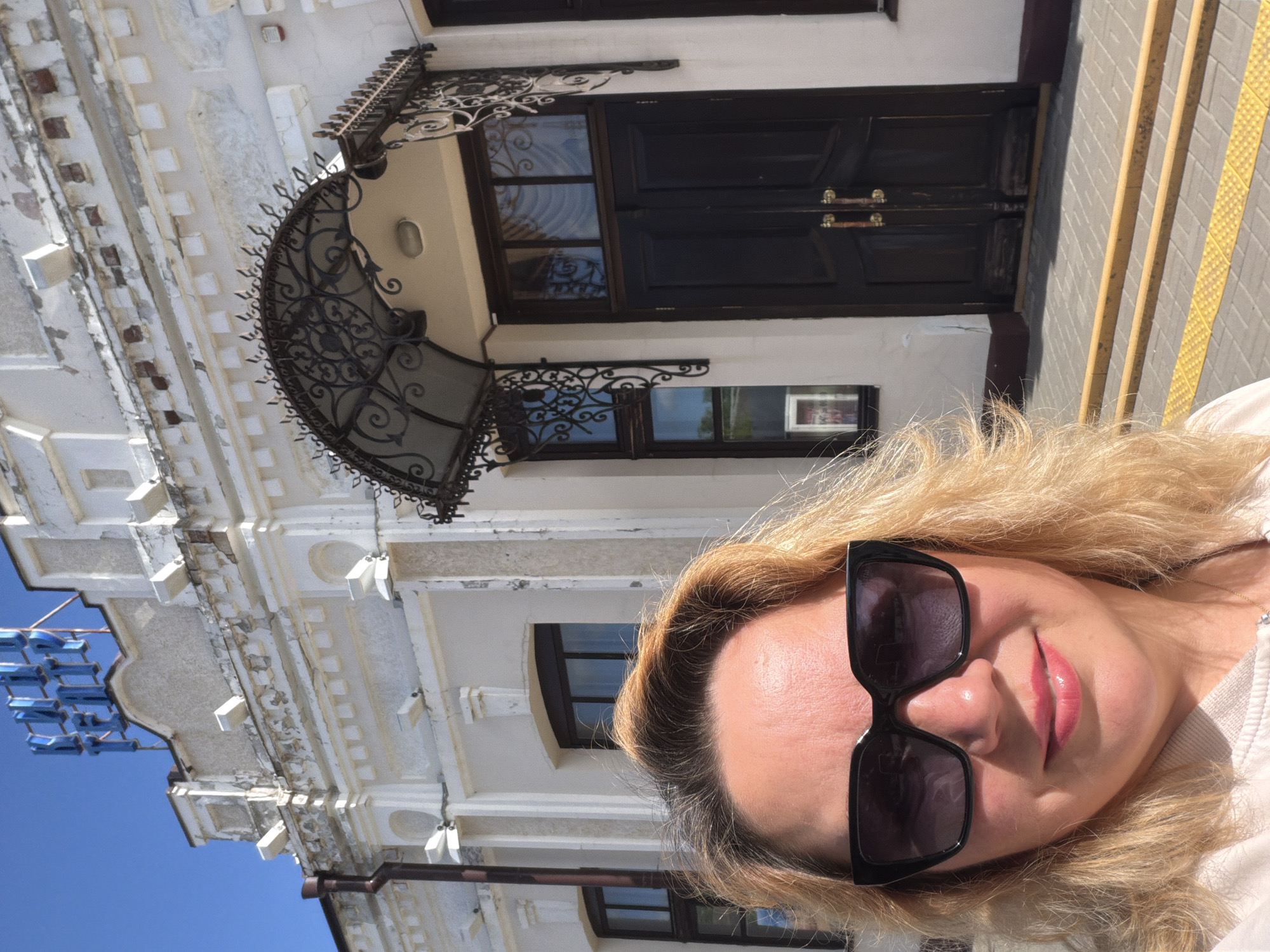 The photograph shows a person with blonde hair and sunglasses, posing in front of a building. The building is white with ornate architectural details, including a decorative metal canopy over the entrance. The entrance consists of dark double doors with metal handles. There are windows visible on the building facade, and a paved area with yellow tactile paving is in the foreground. The picture was taken in Leningradskaya, Russia.