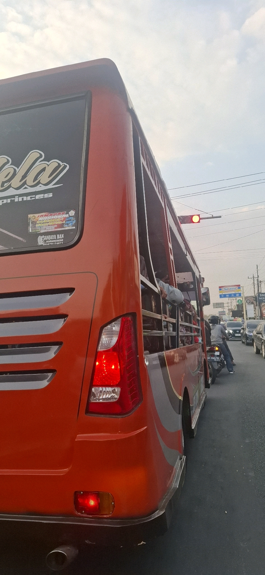An exterior, low-angle, full-shot photograph depicts a bright orange, open-air bus in motion, seen from its rear left quarter, on a paved road. The bus's rear taillight assembly is illuminated red, indicating its brake lights are on. Several people are visible within the bus, with some leaning out of the open sides. The bus features decorative decals on its rear window, one with the word "Princes" and another containing text and logos, including "SANJAYA BAN KARANGGEDE". In the background, a traffic light is red, and several vehicles, including cars and motorcycles, are also present on the road. The sky above is overcast with visible clouds. The environment appears to be an urban or suburban setting in Desa Ngesrep, Indonesia, with buildings and signs faintly visible in the distance, suggesting a commercial area.