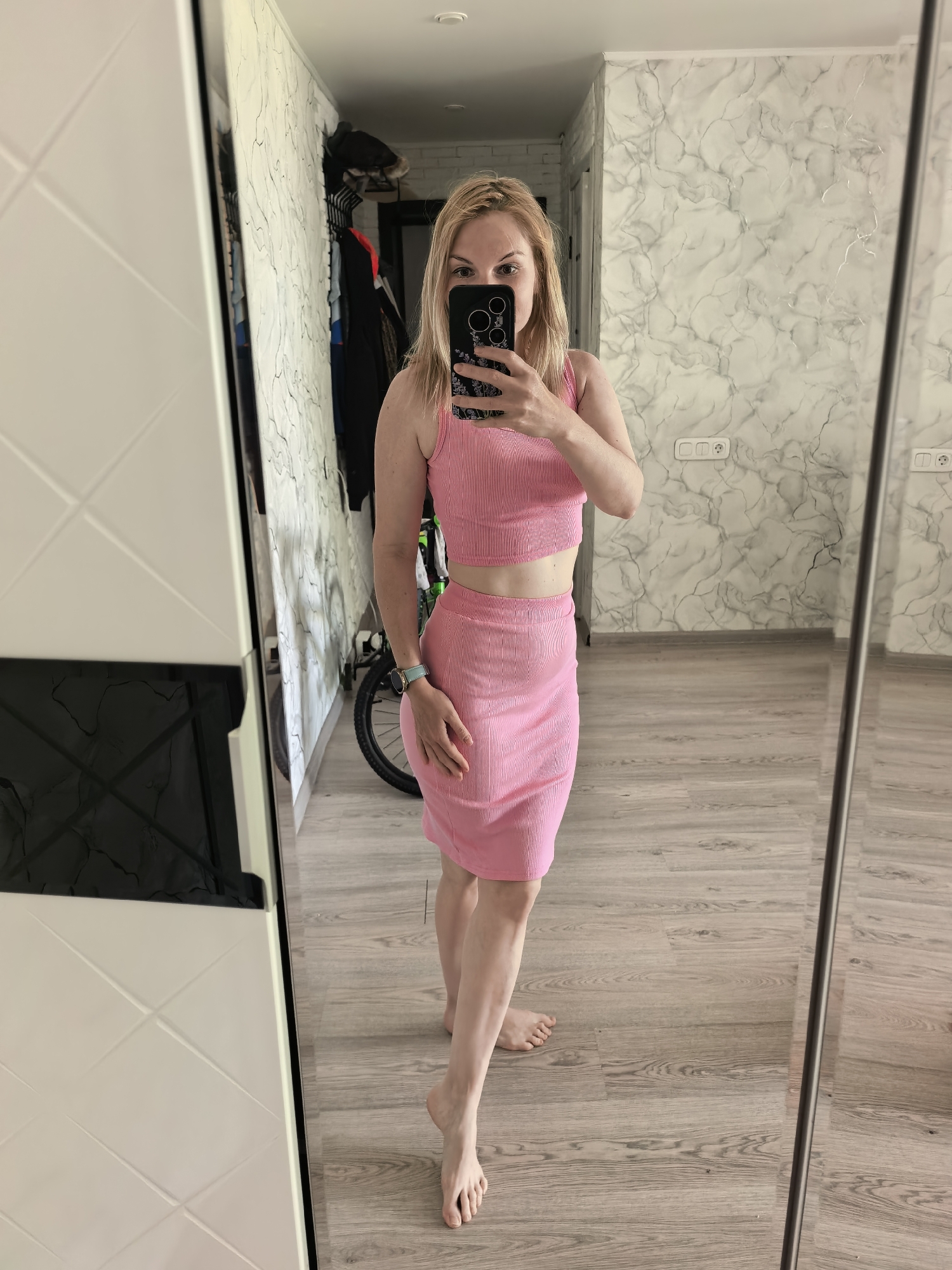 A full shot of a woman with blonde hair wearing a pink two-piece outfit consisting of a cropped tank top and a knee-length skirt is reflected in a full-length mirror. She is holding a smartphone up to take a selfie, obscuring part of her face. She is standing barefoot on a wooden floor, likely in a hallway or room with light-colored, marble-patterned wallpaper. Behind her, visible in the mirror, is a coat rack with clothes hanging on it and part of a bicycle. The woman is wearing a watch on her left wrist. The location could be an apartment or house in Sterlitamak, Russia.