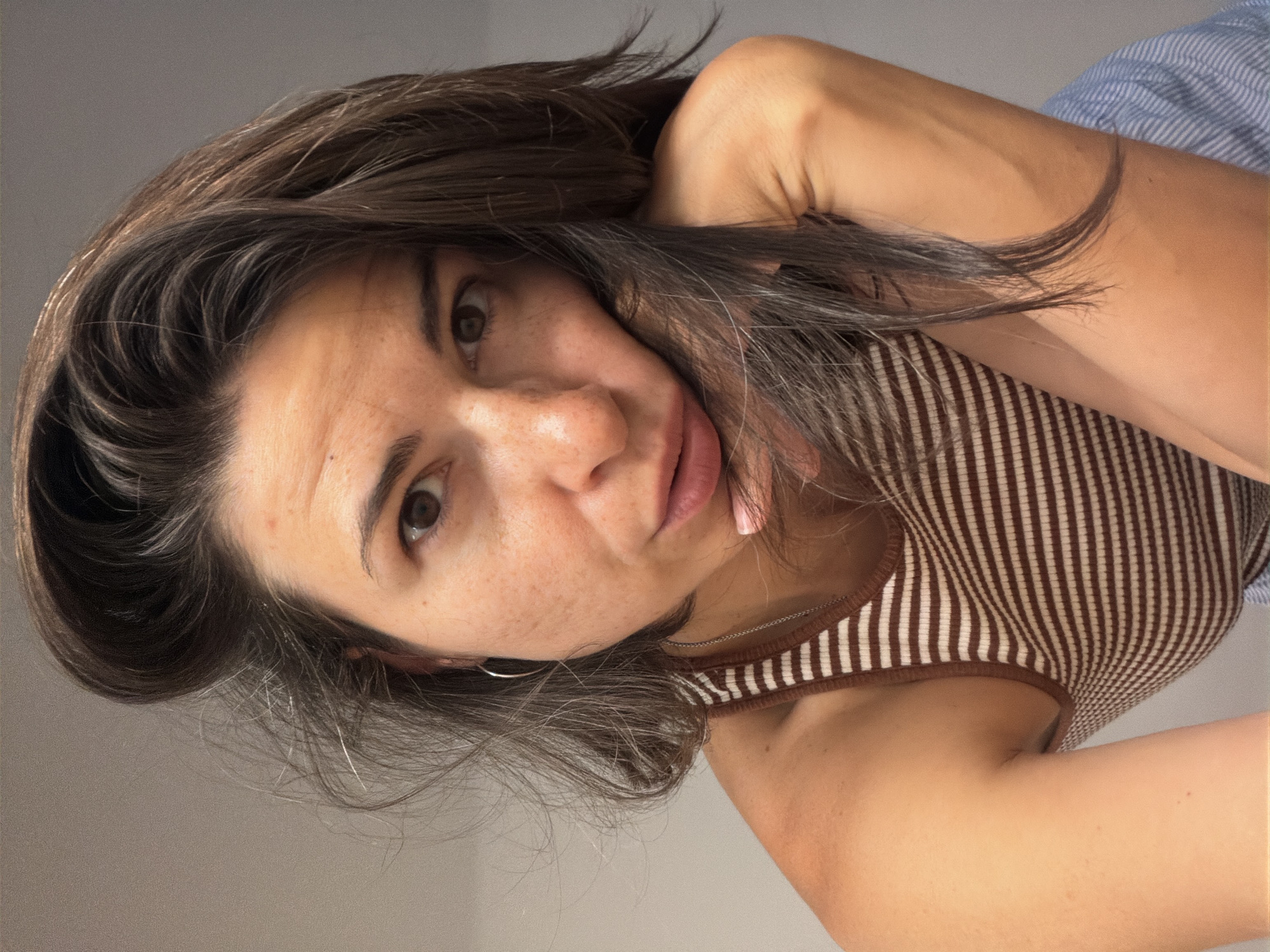 A woman with light skin, brown hair, and brown eyes looks at the camera. She has freckles on her face and is wearing a striped brown and white tank top. Her left arm is near her head. The background appears to be a light grey wall. The scene does not feature Budva, Montenegro.