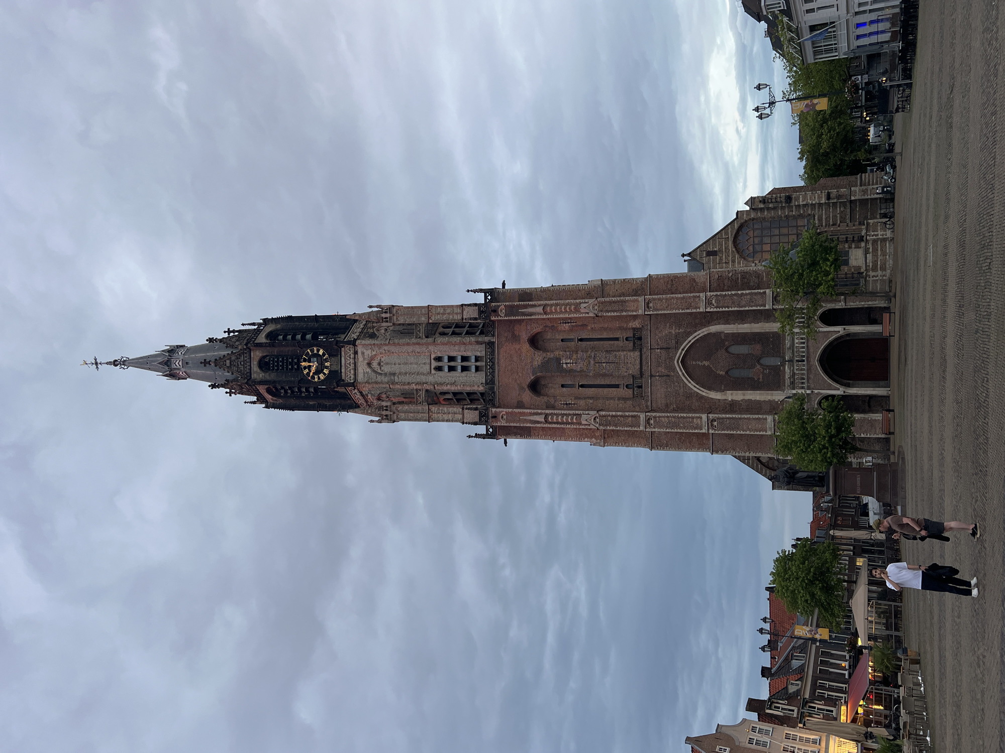 The image captures a wide view of the Markt square in Delft, Netherlands, dominated by the imposing Oude Kerk (Old Church). The Oude Kerk's tall, brick tower, featuring a clock face and a spire, rises towards a cloudy, overcast sky. The lower portion of the church is visible, showing arched doorways and windows.
In the foreground, a paved square is sparsely populated. Two men are standing and conversing in the center of the square. One man, wearing a white t-shirt and dark pants, holds a black bag. The other man, in a brown t-shirt and dark shorts, also holds a black bag.
To the left of the square, a row of historic buildings with red-tiled roofs line the street. Some of these buildings appear to be shops or restaurants, with awnings and outdoor seating visible. Trees are scattered throughout the scene, adding greenery. A statue on a pedestal is situated near the church.
To the right, more buildings and streetlights are visible. A scooter is parked near the church. The overall atmosphere suggests a late afternoon or early evening with diffused light due to the cloud cover.