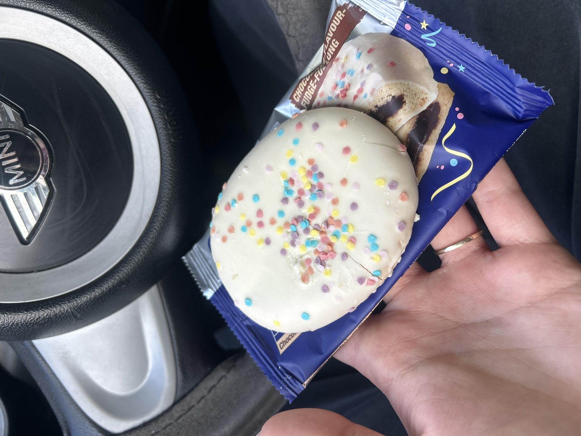 The image shows a hand holding a packaged cake next to a Mini Cooper steering wheel. The cake is round, covered in white frosting, and sprinkled with colorful sprinkles. The packaging is blue and features an image of a chocolate fudge-filled cake. The hand has a wedding ring on the ring finger. The steering wheel is black with the Mini Cooper logo visible. The location cannot be determined to be Burgas, Bulgaria, based on the visual information provided.