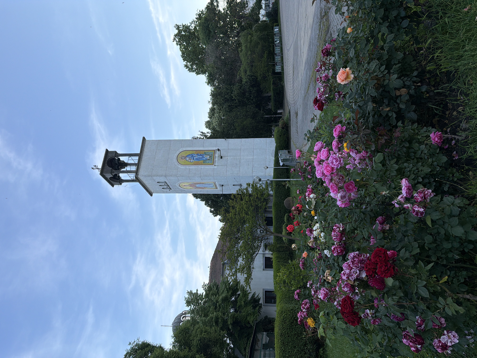 The image displays an outdoor scene in Burgas, Bulgaria, featuring a church complex with a prominent bell tower and a rose garden in the foreground under a light blue, partly cloudy sky.
In the center-right, a tall, white rectangular bell tower stands vertically. Its visible face features two arched mosaic icons, each depicting a robed saint; the left icon shows a figure holding a book, and the right a similar figure, both in blue and gold tones. A small black camera or sensor is mounted on the left side of the tower, below the left mosaic. The top section of the tower consists of an open belfry structure with dark, arched openings supported by columns, surmounted by a dark cross. To the left of the bell tower, partially obscured by green trees, a white building with a dark, domed roof and a small cross at its apex is visible, indicative of a church. Another building with a terracotta-tiled roof is also visible behind the trees.
The foreground is dominated by a dense, vibrant rose garden featuring numerous rose bushes in bloom. The roses exhibit a variety of colors including deep red, various shades of pink (fuchsia, light pink), mottled purple-pink, yellow, and peach/orange. The foliage of the rose bushes is dark green. Below the rose garden, green grass is visible.
Surrounding the buildings and the rose garden are numerous mature trees with dense green foliage, creating a lush environment. Hedges are also present, forming boundaries. To the right of the rose garden and extending into the midground, a wide, patterned paved area, likely cobblestone or pavers, forms an open plaza or pathway. Along the far right edge of this paved area, several low, dark rectangular structures (possibly display boards or planters) are arranged. The overall lighting suggests either early morning or late afternoon, with soft, diffused light. No people are visible in the scene. Small, faint bird-like specks are discernible against the sky.