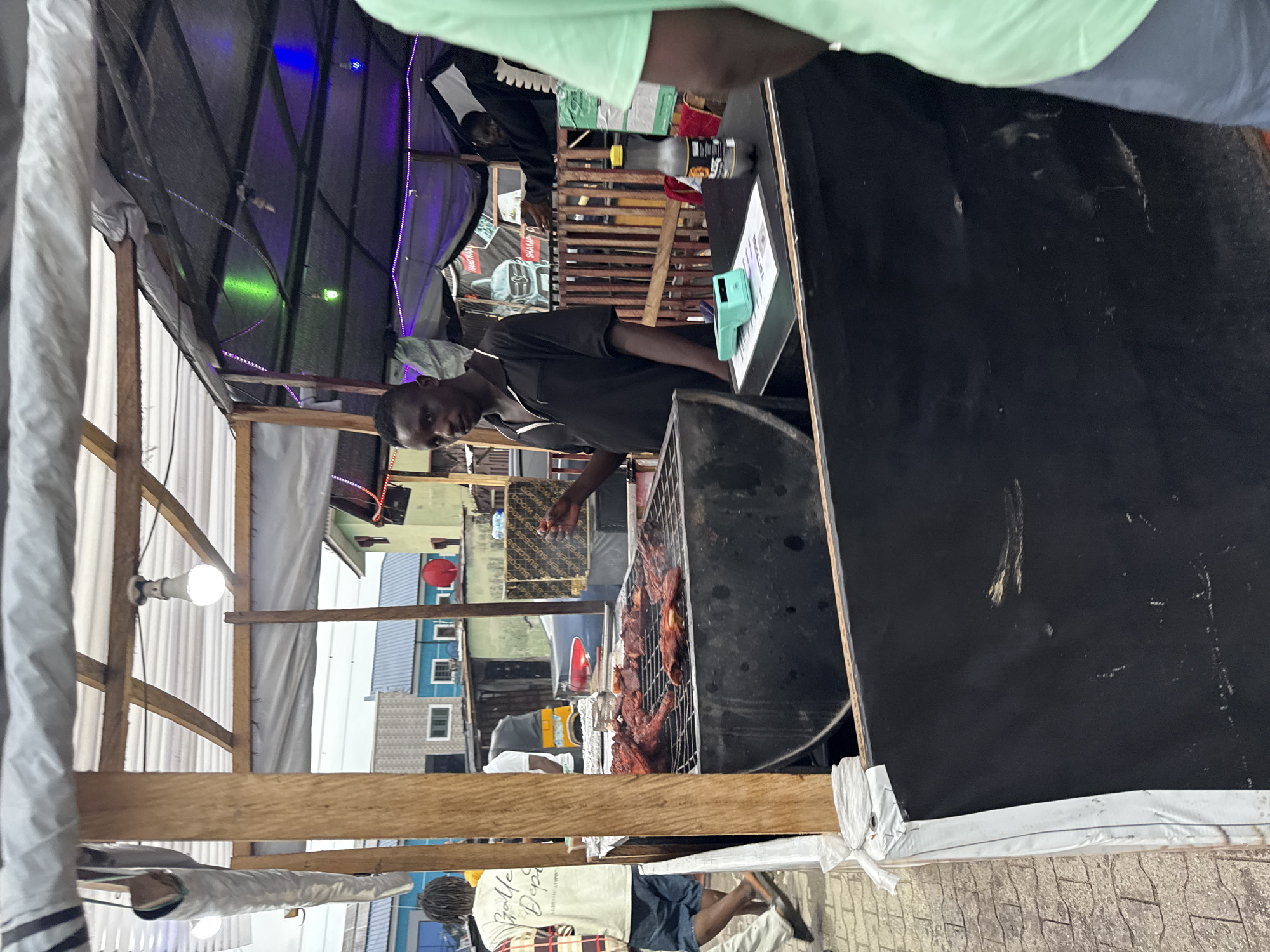The image shows a man grilling meat at an outdoor food stall in Ado, Nigeria. He is standing next to a charcoal grill laden with cooking meat. The stall is constructed with wooden beams and covered with tarp, lit with LED string lights. A menu display and a payment terminal are visible on the counter. In the background, a street scene is visible, with residential buildings and a commercial establishment. People walk around the stall, some seemingly customers.