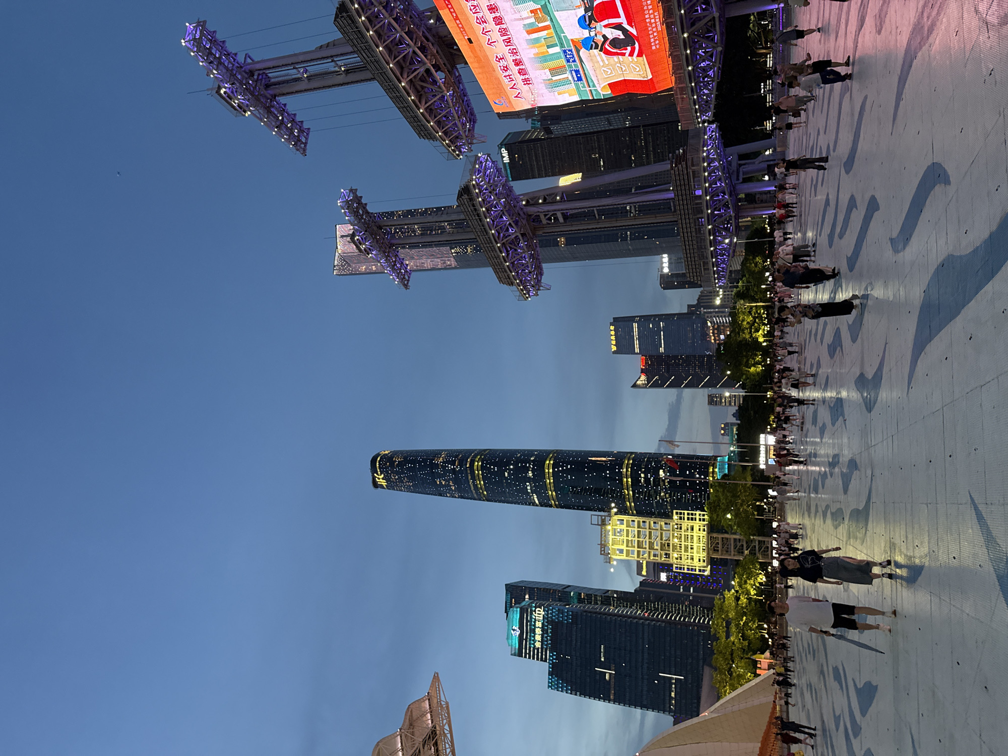 This image depicts a wide, paved public plaza at dusk in Tianhe District, Guangzhou, China. In the foreground, several individuals are walking across the plaza, which features a light blue surface with abstract, darker blue patterns creating shadows. To the left, a large, white, conical structure is partially visible. Dominating the background are several modern skyscrapers, illuminated with various lights. The tallest building, a sleek, dark tower with gold and white lights, is prominently featured. To the right, a large LED screen displays text and graphics in Chinese, with a portion of a red banner visible. Above the plaza, elevated structures with purple lighting are visible, possibly part of an entertainment complex or observation decks. The sky is a deep blue, transitioning from daylight to twilight. The overall scene suggests a bustling urban environment at the end of the day, with people enjoying the public space and the illuminated cityscape.