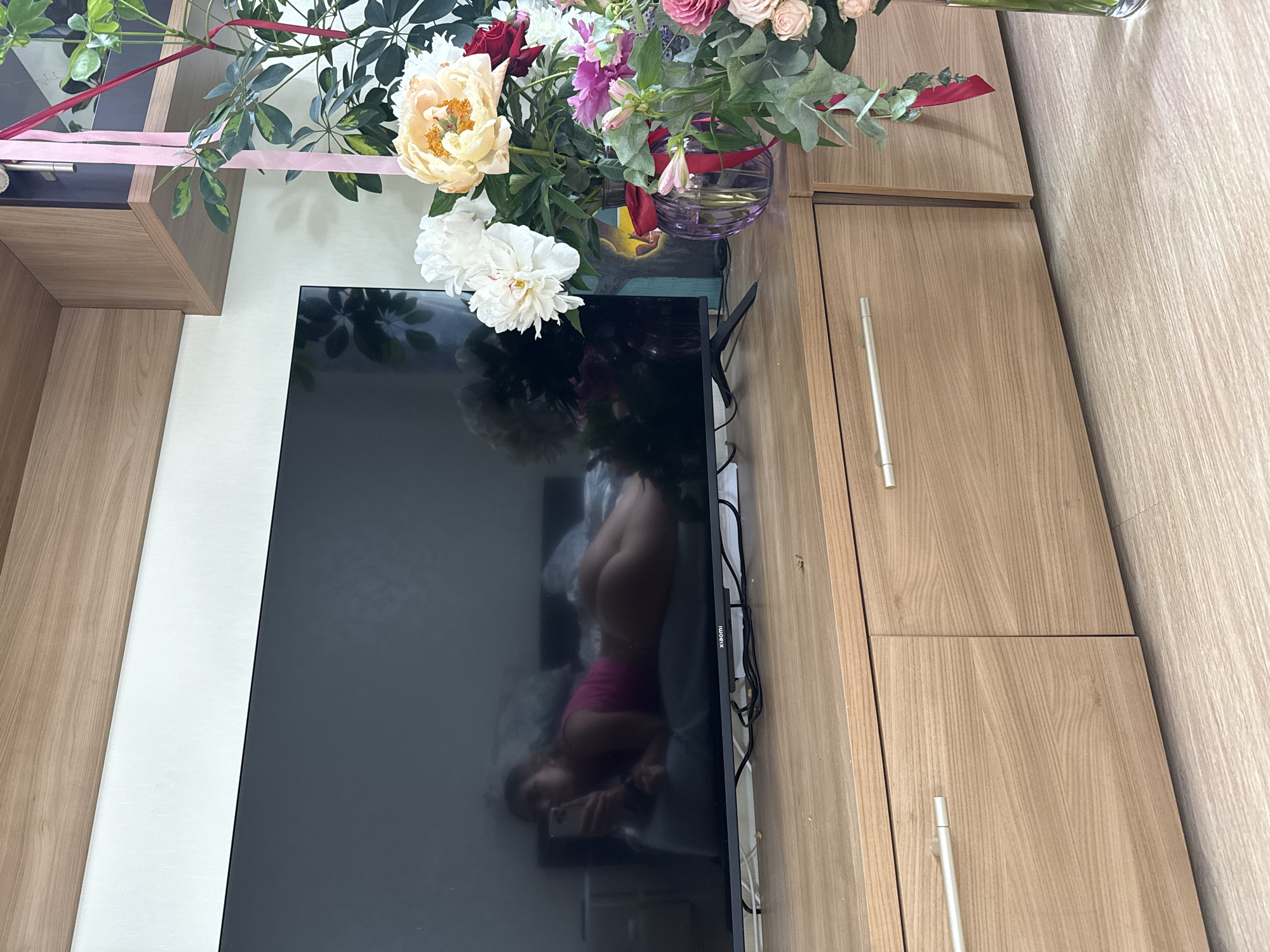 The image shows a still life arrangement of flowers and a television. A large bouquet of various flowers, including peonies, roses, and alstroemerias, is arranged in a glass vase with a red ribbon. The vase sits on a glass surface, possibly a table or shelf, next to a black flat-screen television. The television screen reflects an image of a person lying down, with their back to the camera, wearing pink shorts. The setting appears to be indoors, with wooden cabinetry visible in the background. The lighting suggests it is daytime. The overall scene is composed and appears to be staged for a photograph. There is text visible on the television's bezel that appears to read "XIAOMI".