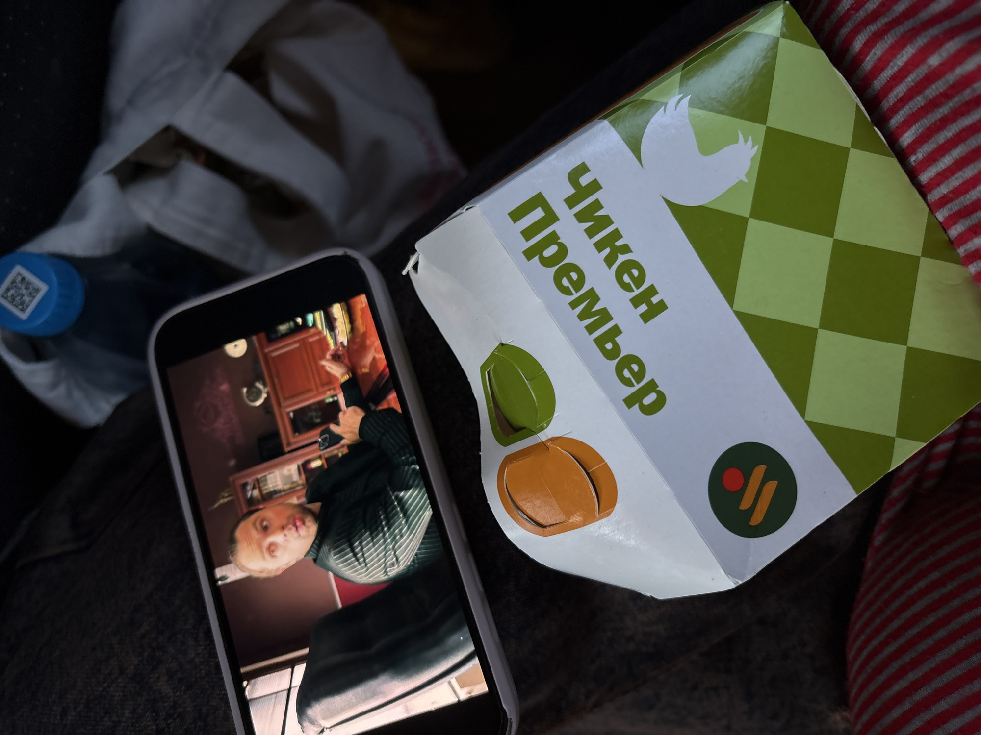 The image shows a person holding a smartphone displaying a video of a man, likely a selfie or live stream, next to a "Chicken Premier" food box written in Cyrillic on what appears to be the interior of a vehicle. There is also a bottle of water and a white cloth bag in the background. There is a red and gray striped seat. The location is likely in Usolye-Sibirskoye, Russia based on the text on the box.