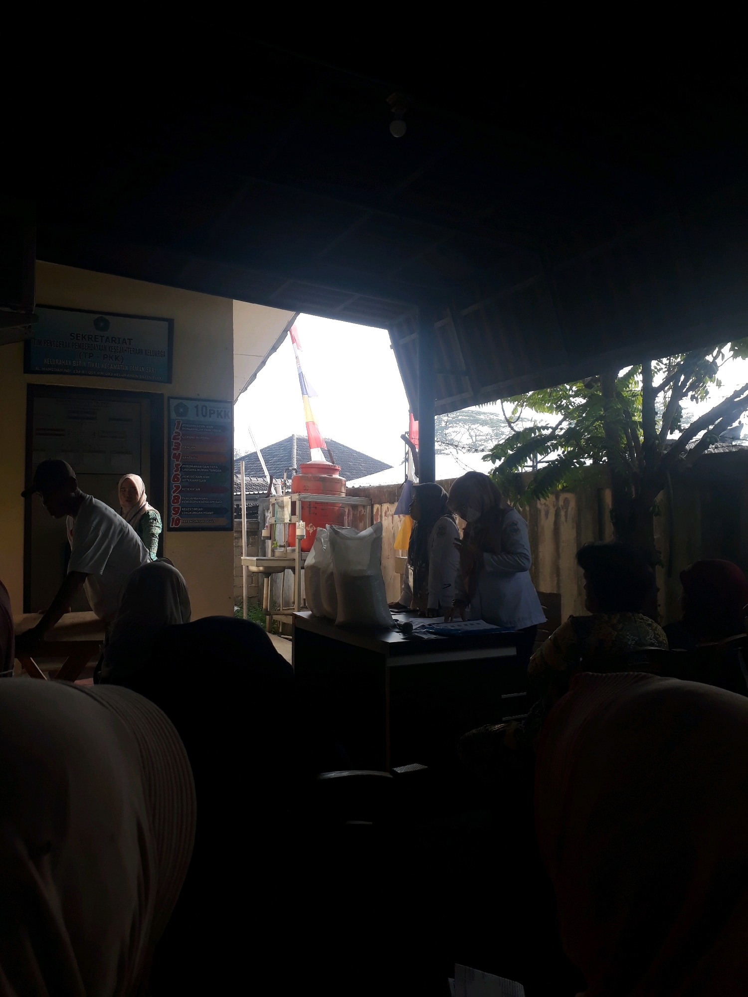 The image shows an outdoor covered area, possibly a community center or office, with several people present. A sign on the wall reads "SEKRETARIAT TIM PENGGERAK PEMBERDAYAAN KESEJAHTERAAN KELUARGA (TP-PKK) KELURAHAN BATIN TIVAL KECAMATAN TAMAN SARI". This indicates the location is the Secretariat of the Family Welfare Empowerment Movement Team in Batin Tival Village, Taman Sari District. There are two women in the foreground wearing light-colored tops and headscarves, appearing to be attending to some papers on a table. A man in a white t-shirt and cap is visible to the left, also near a table. In the background, a large red water tank with a colorful flag on top is visible, along with several bags of what appears to be white powder. The overall lighting suggests it is daytime, with bright light coming from the sky, possibly indicating an overcast or hazy condition. Trees and a building with a tiled roof are also visible in the background.