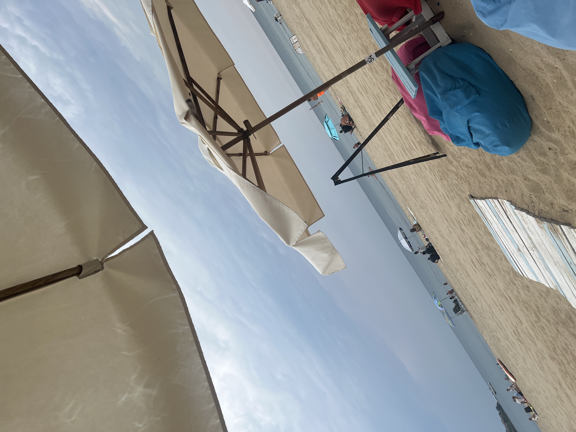 This image depicts a cloudy day at a sandy beach in Sozopol, Bulgaria. In the foreground, two large beach umbrellas are visible, one dominating the left side of the frame and another in the mid-right. Underneath the second umbrella, there are beanbag chairs in pink and blue, and a small blue table with the number "54" on it. A wooden boardwalk with blue and white planks is partially visible in the bottom left corner.
Further out on the beach, several people are scattered, some sitting under umbrellas, others walking or standing. The sea stretches to the horizon under a gray, overcast sky. A few boats are visible on the water in the distance. The overall scene suggests a typical beach day with people enjoying the seaside amenities.