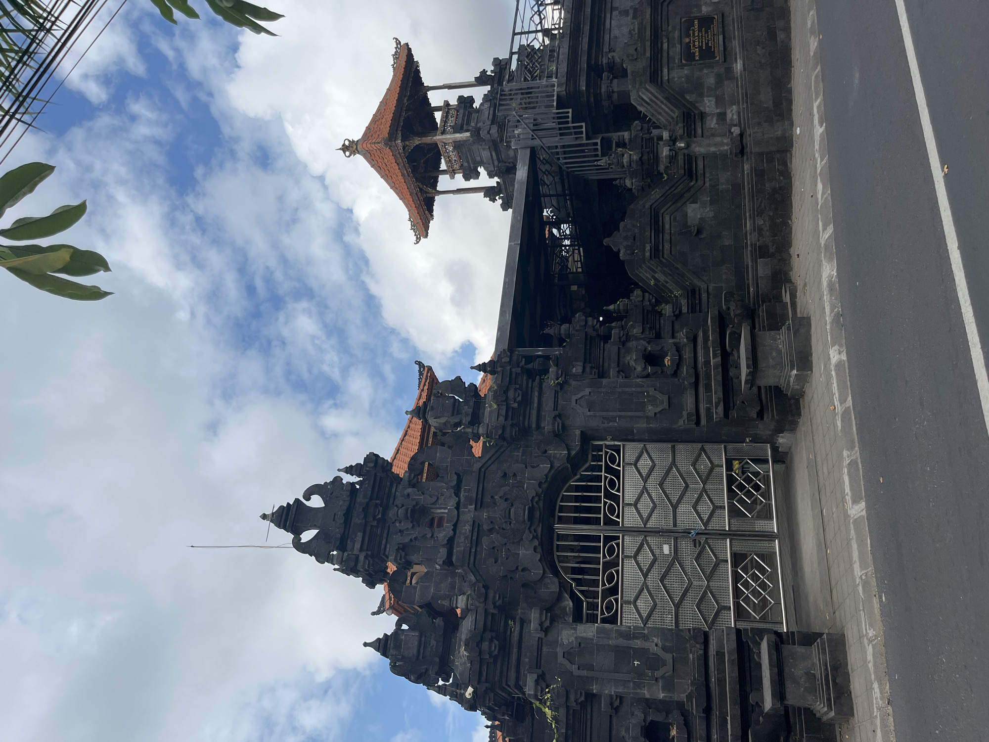 This is a low-angle, medium shot of a Balinese temple gate in Kuta Selatan, Indonesia. The temple gate is constructed from dark, carved stone and features intricate details. A large, silver metal gate with a geometric pattern is visible in the center. To the right of the main gate, a smaller, multi-tiered structure with a reddish-orange tiled roof and wooden railings is visible. A staircase leads up to this structure. The sky above is filled with white, fluffy clouds against a blue backdrop. Green foliage is visible in the upper right and upper left corners of the image, suggesting the presence of trees or other plants nearby. The foreground shows a paved sidewalk and a gray asphalt road with a white line marking. There are no people or animals visible in the scene. The overall impression is one of a significant religious or cultural landmark.