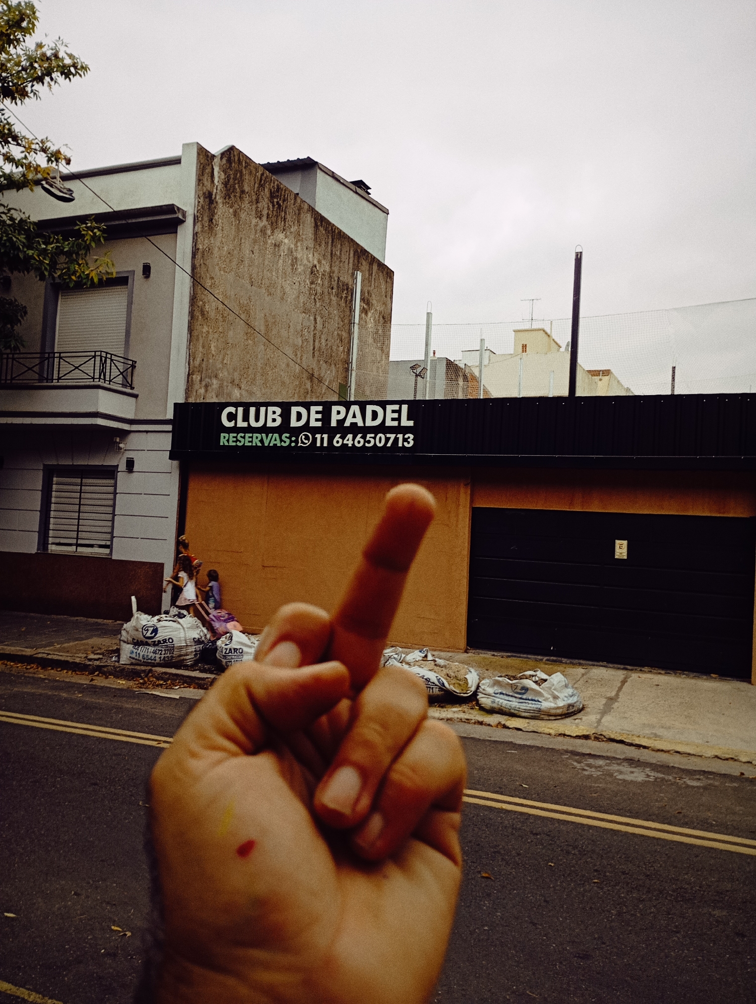 The image shows a hand giving the middle finger in front of "Club de Padel" (Padel Club) with the phone number for reservations indicated. The club is located on a street in Buenos Aires, Argentina. Two children and an adult are seen near a pile of filled white bags in front of the building. The bags have a blue and white design. There are two yellow lines on the road in front of the Club. There's a building on the left with a small balcony. The sky is overcast.