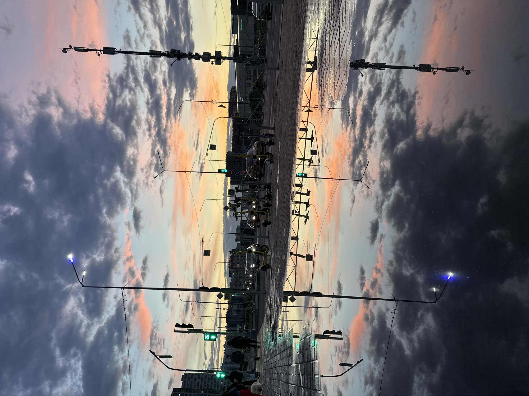 A photograph taken at ground level shows a large puddle reflecting a city street scene at dusk. The sky is filled with dramatic clouds in shades of gray, pink, and orange, with the last light of sunset visible on the horizon. Several streetlights are visible, some with illuminated bulbs casting a blue and white light. A tall cell tower stands on the right side of the frame, its structure and antennas clearly defined in the reflection.
On the street, there are several parked electric scooters, some of which are yellow. Cars are visible driving or parked in the background, their headlights on. Pedestrians walk along the sidewalk on the left side of the image, one of whom is walking a white dog. Traffic lights are also present, with one showing a green light. The wet pavement and the large puddle create a mirror-like surface that reflects the sky, the streetlights, the cell tower, and some of the surrounding structures. The overall impression is a moody, atmospheric urban landscape at the end of the day.