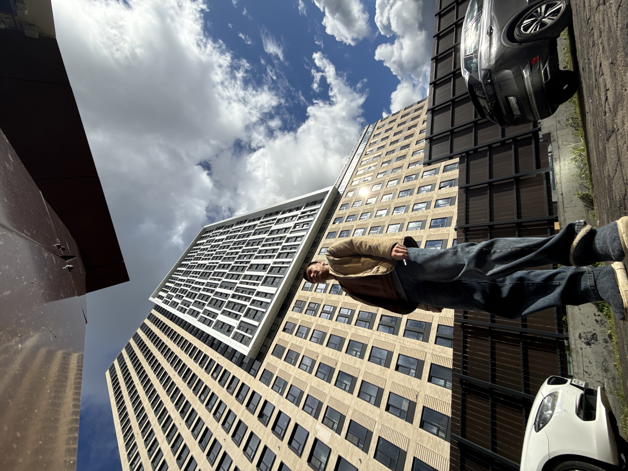 A low-angle perspective shows a man standing on a sidewalk adjacent to tall buildings under a partly cloudy sky. The location is Khimki, Russia. The man is wearing a brown jacket and blue jeans and is partially obscured by one of the buildings. To his right, two cars (a gray one and a white one) are parked on the street. The buildings have many windows and a light beige façade. A section of one building has a white exterior with black window frames.