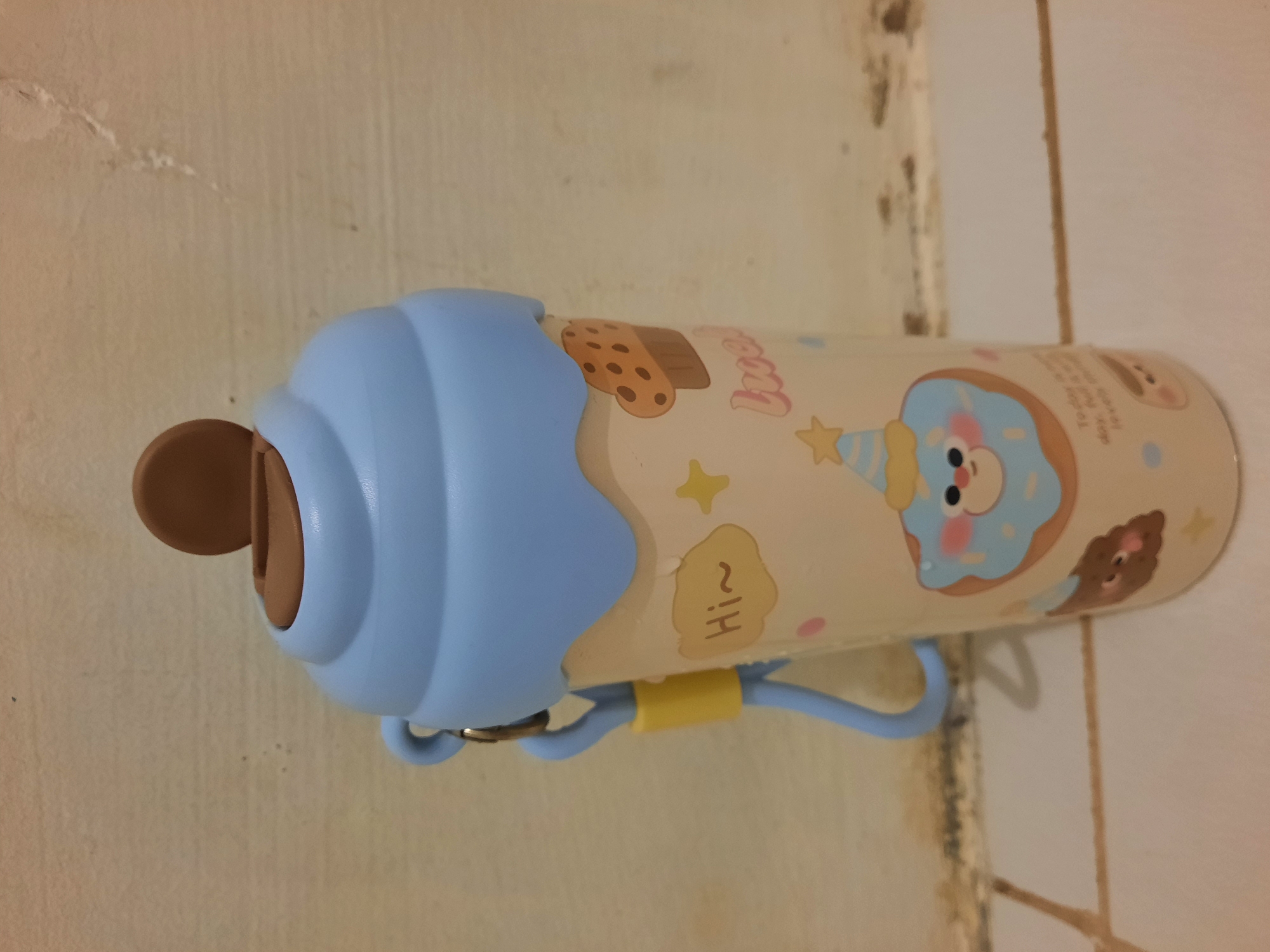 An ice cream-themed water bottle sits on a tiled surface against a textured, light-colored wall. The bottle has a light blue, ruffled lid resembling whipped cream, topped with a brown, disc-shaped cap. A light blue strap with a yellow holder is attached to the side of the lid. The bottle's body is cream-colored and decorated with various cute illustrations, including a smiling blue donut wearing a party hat, a cupcake, stars, and polka dots. Text is visible on the lower part of the bottle, reading "Hi~" and "Today is a lovely day, hope its lovely too". The lighting suggests an indoor environment, possibly a home setting. The scene is located in Lhokseumawe, Indonesia.